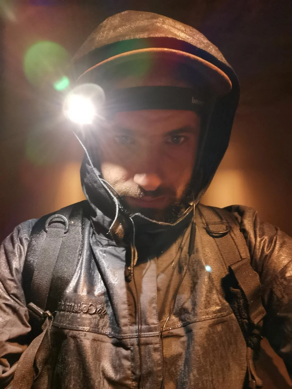 A man wearing a helmet with a headlamp and a waterproof jacket, carrying a backpack, taking a selfie in a dark environment.