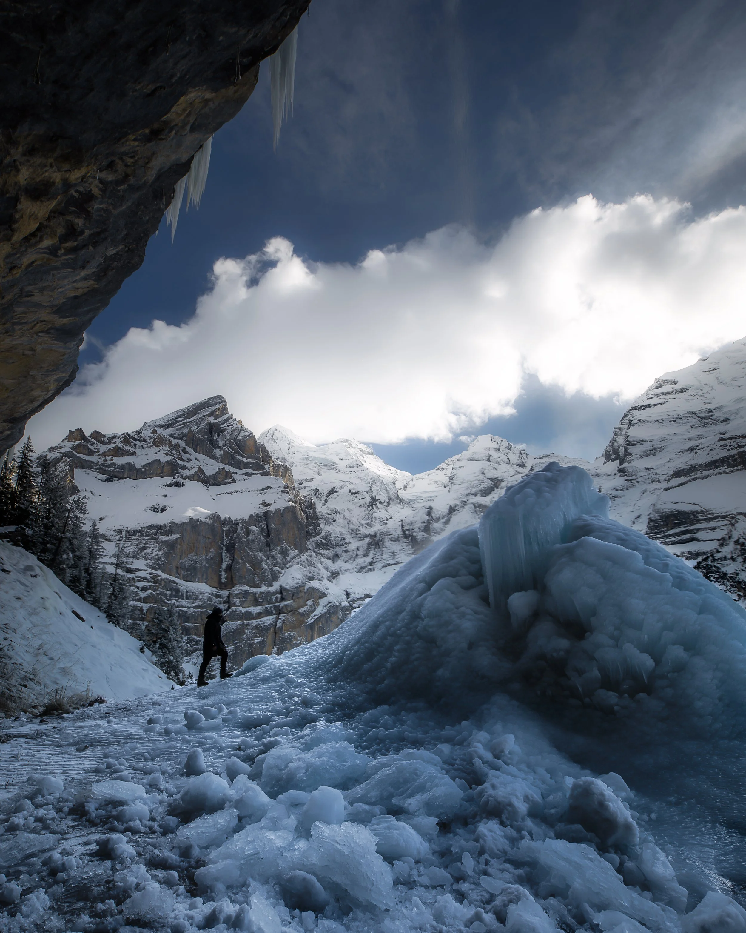 Snow-covered mountain landscape with a person standing on the icy ground and large ice formations in the foreground, tall snow-capped peaks in the background, and a partly cloudy sky overhead.