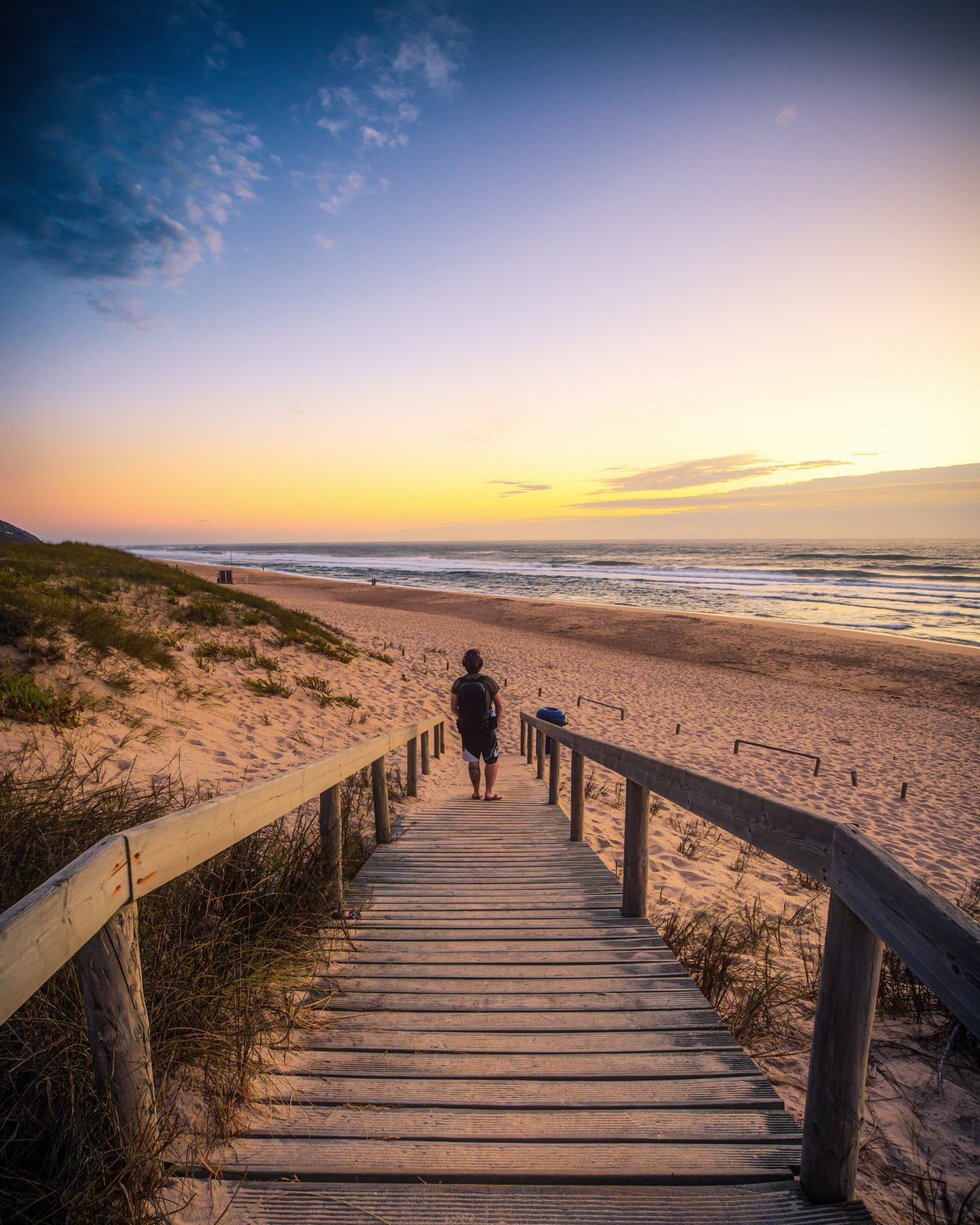 A person walking down a wooden boardwalk towards a beach at sunset, with the ocean and sky in the background.