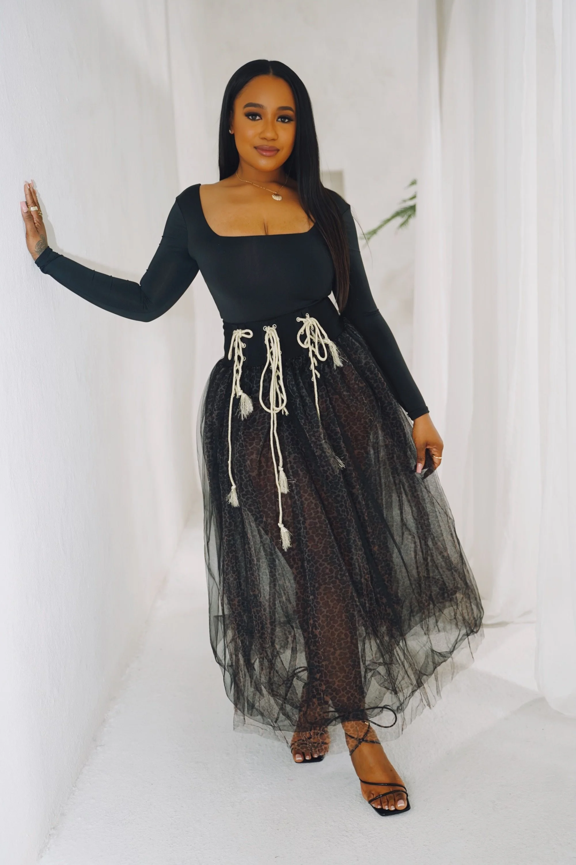 A woman in a black long-sleeve top and a sheer black skirt with a leopard print pattern, standing in a white room, posing with one hand on the wall.
