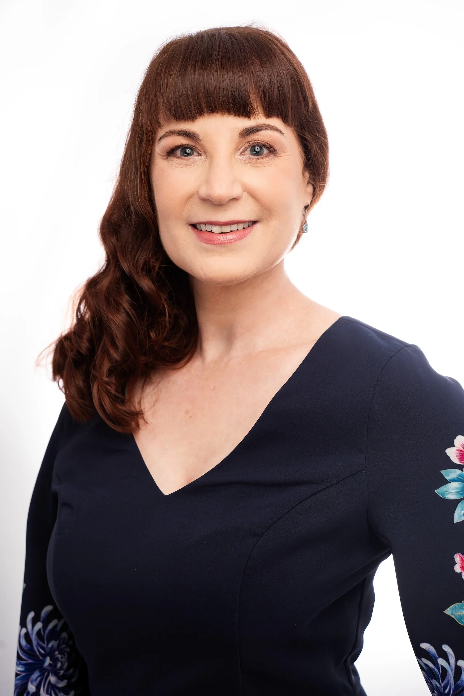 Headshot of a woman with red hair, smiling, wearing a navy-blue top with floral embroidery on the sleeves, against a white background.