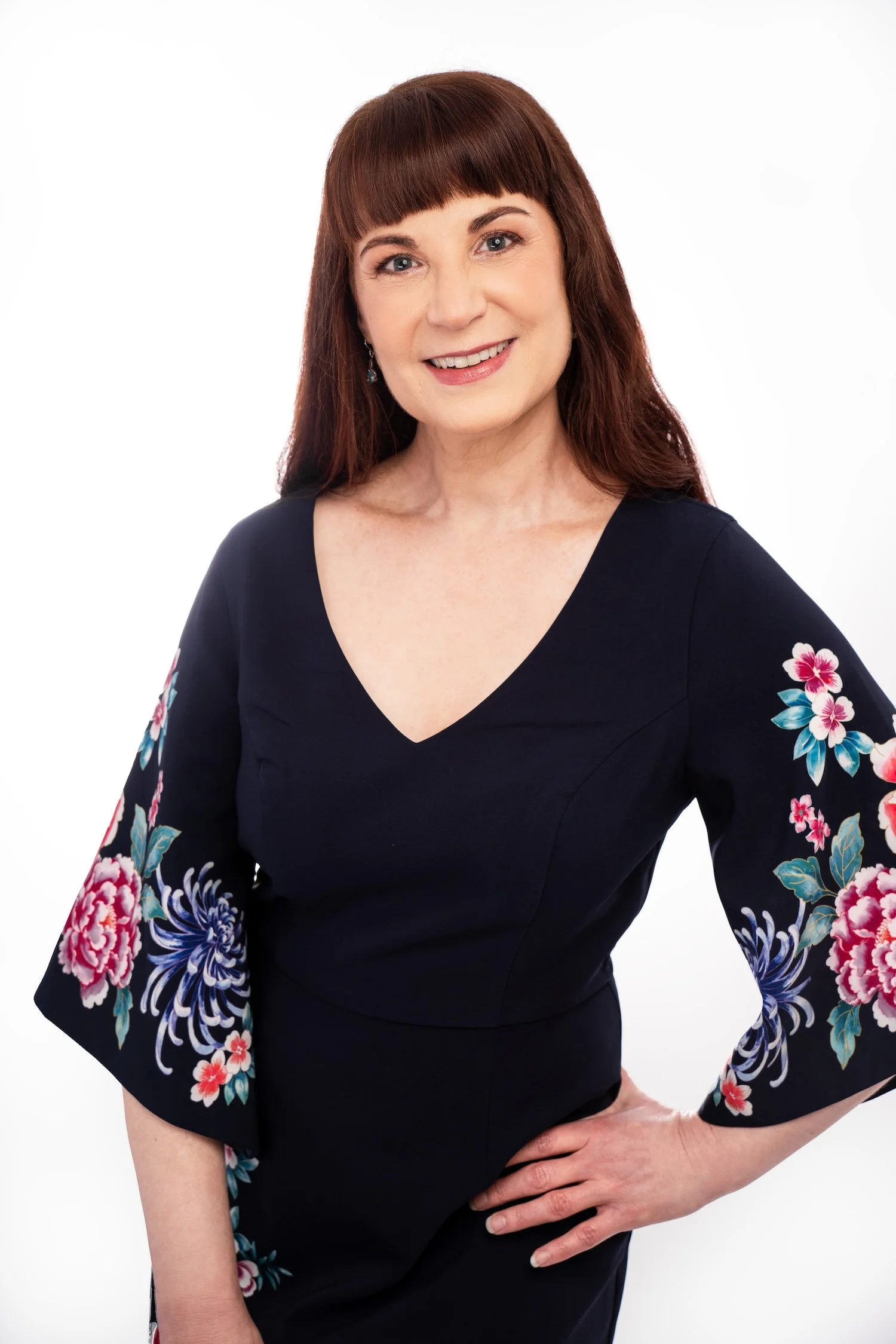 Woman with auburn hair and bangs wearing a navy dress with floral embroidery on the sleeves, smiling at the camera.