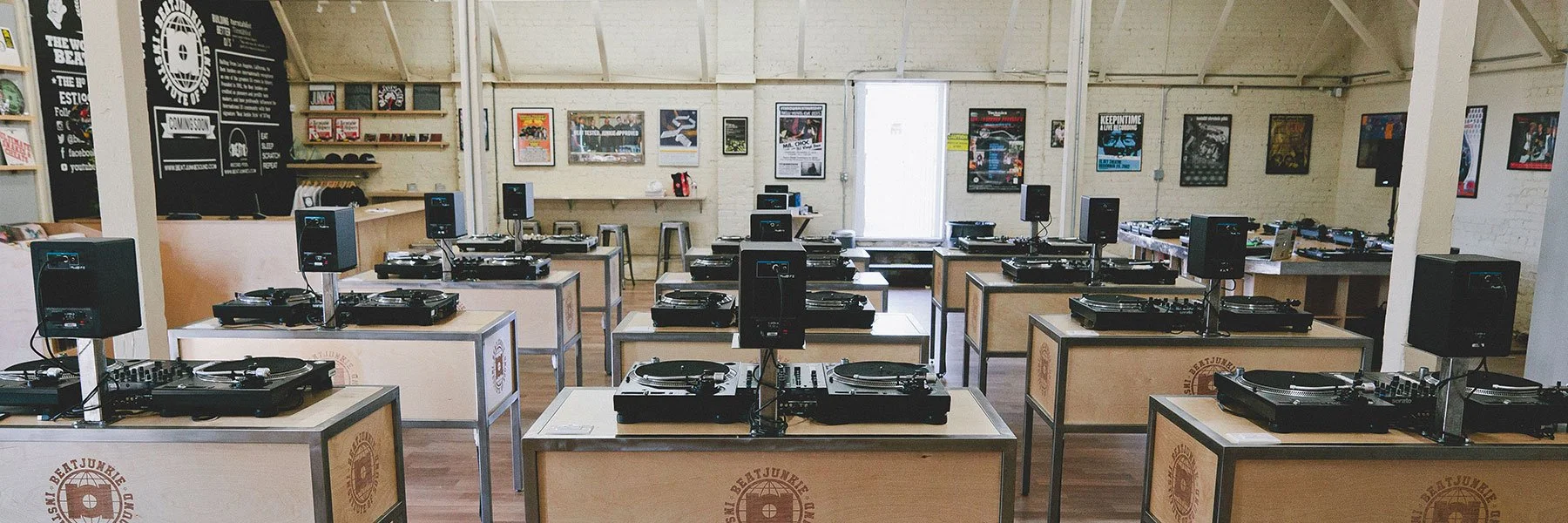 A room set up with multiple DJ decks and speakers on tables, with posters on the walls and a door at the back.