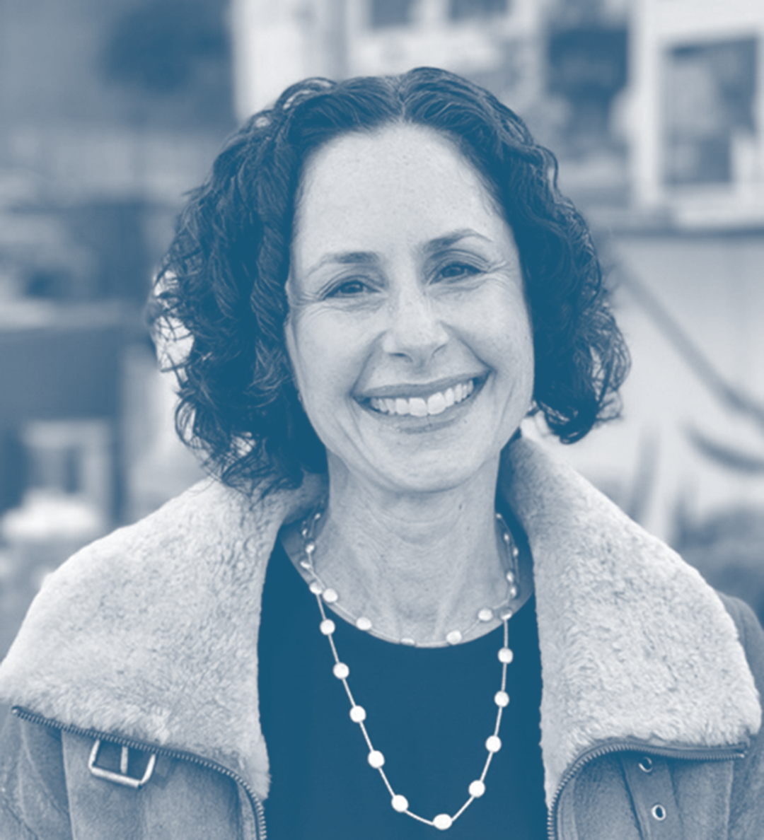A woman with curly dark hair smiling outdoors, wearing a jacket with a fur collar and a beaded necklace.