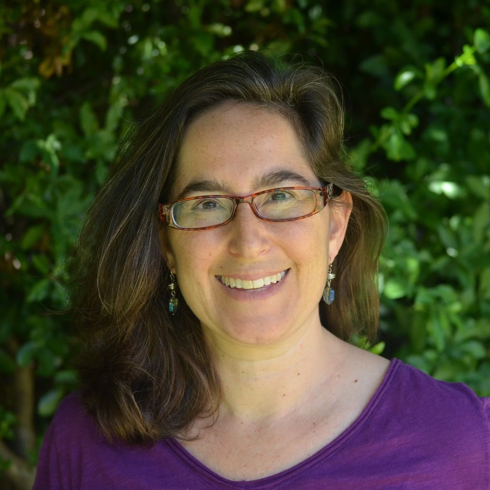 A woman with long brown hair, wearing glasses and dangle earrings, is smiling outdoors with green foliage in the background.