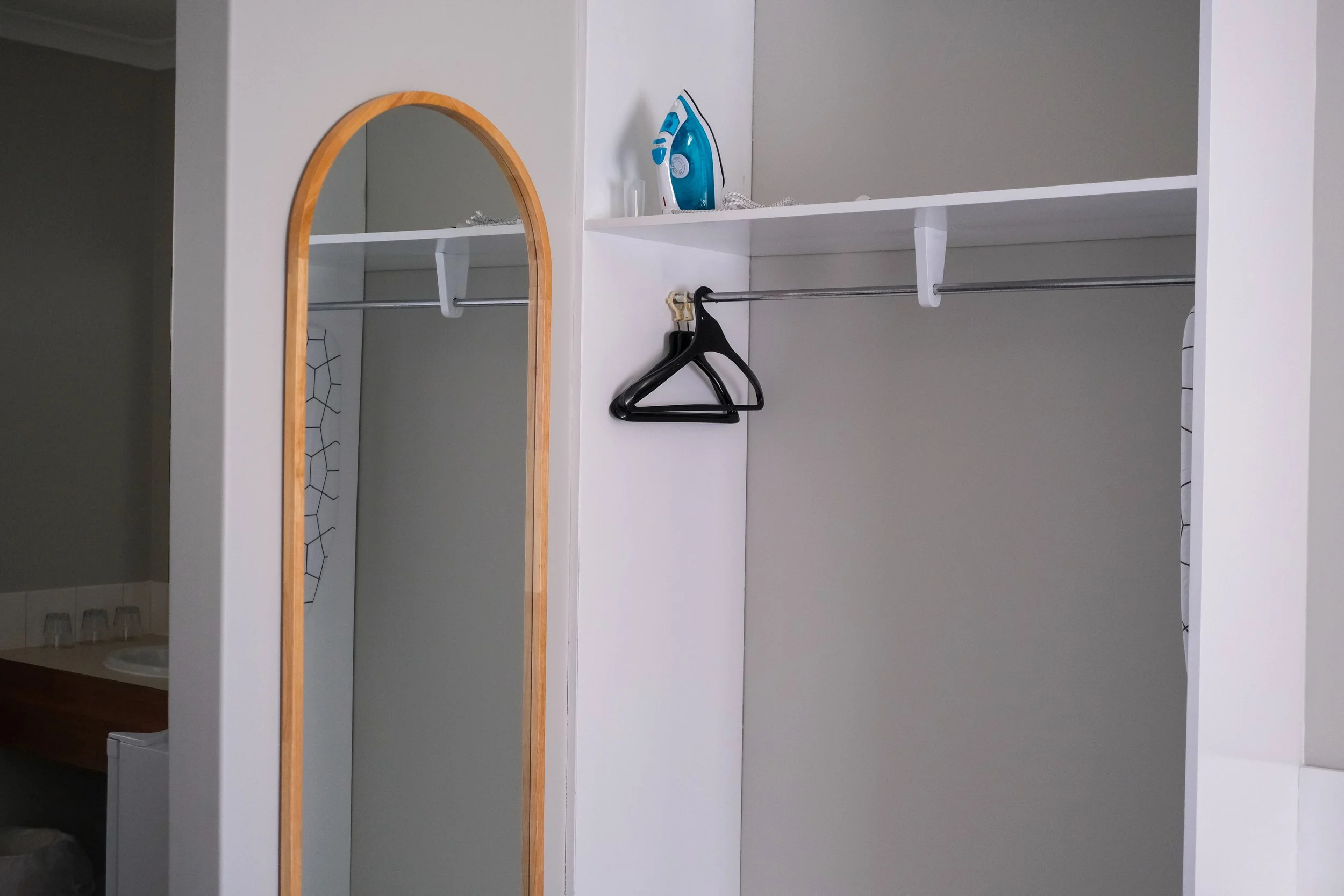 Empty closet with a mirror and an iron on top shelf, some hangers, and a partial view of a bathroom with a sink and small glasses.