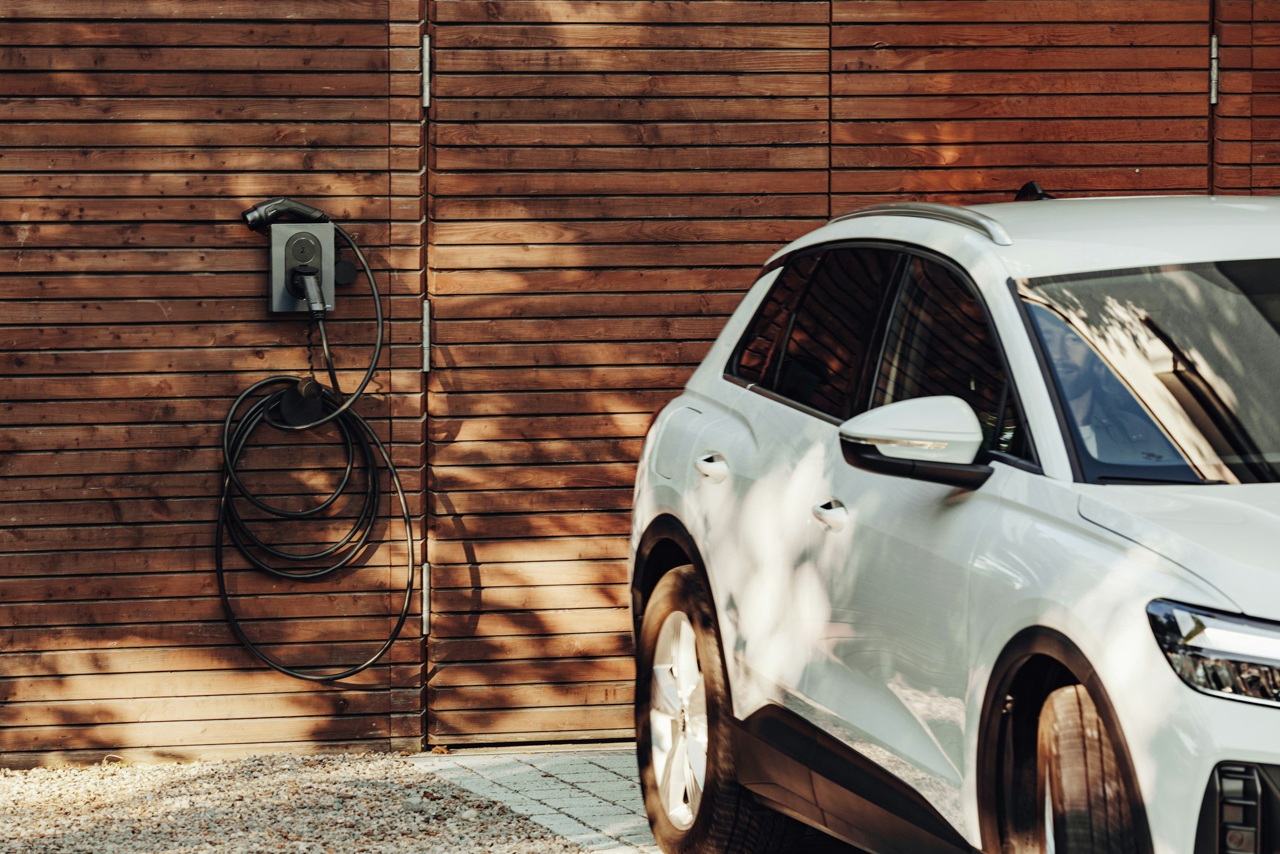 Electric car charging station attached to a wooden garage wall, with a white electric vehicle parked in front.