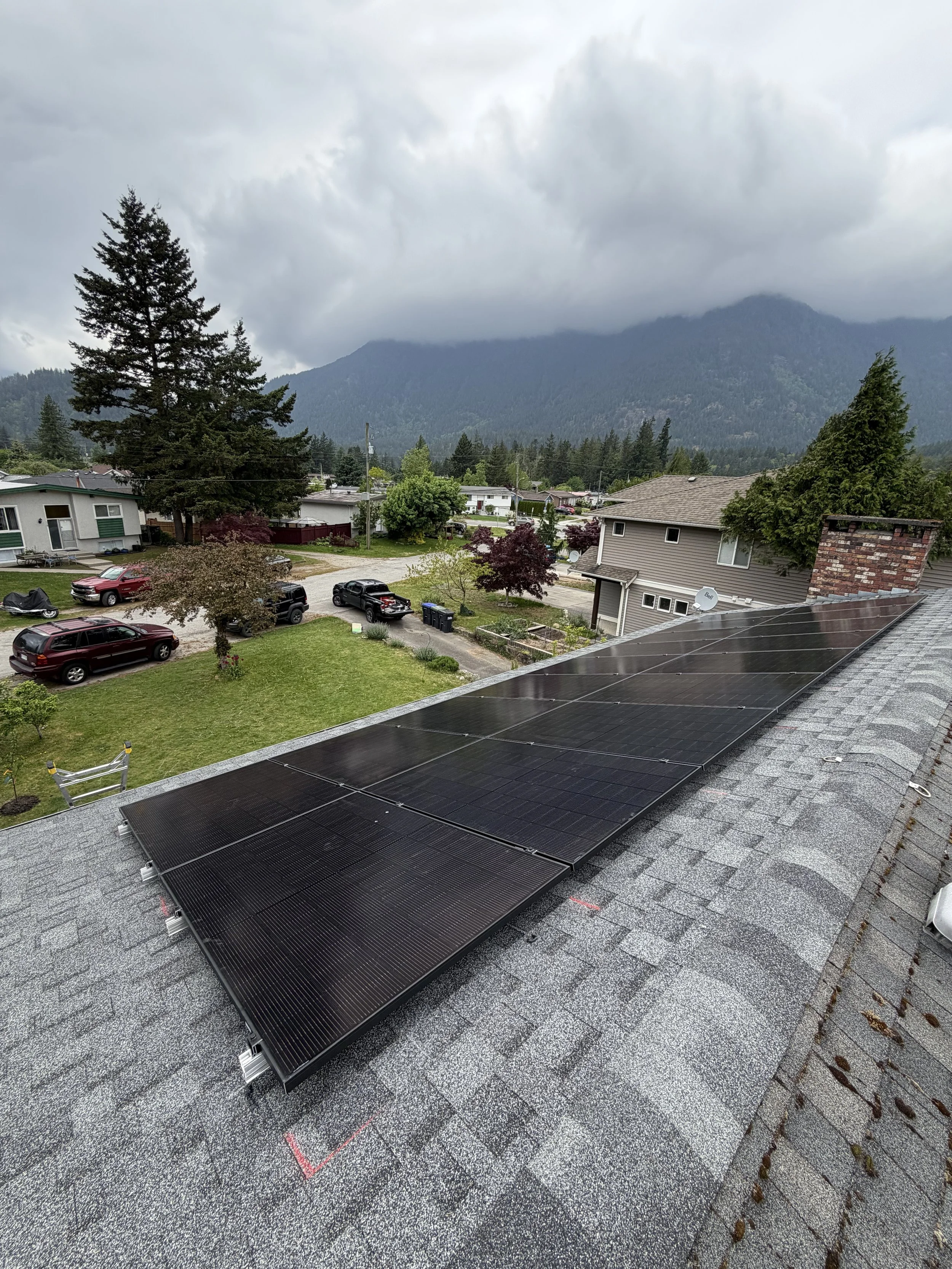 Solar panels installed on a gray shingled roof in a suburban neighborhood with mountains, trees, and houses in the background.