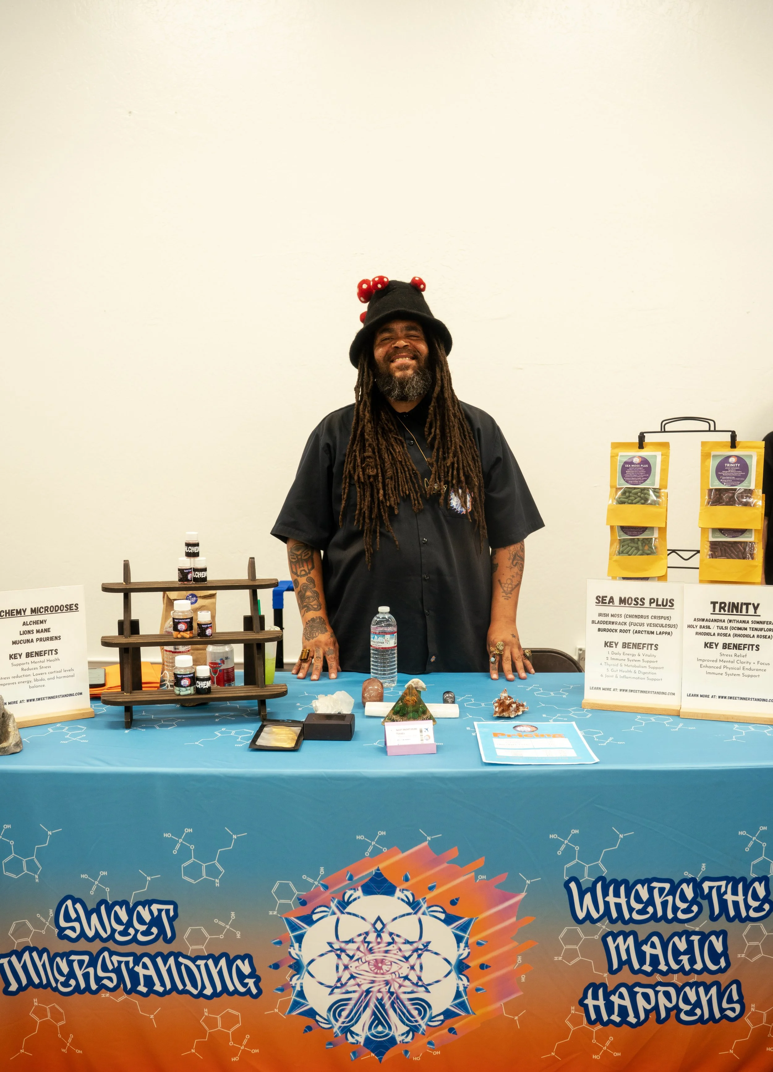 A man with long dreadlocks and a beard smiling stands behind a table with a blue tablecloth at an exhibit booth. The booth has colorful posters with scientific information and displays of natural products labeled Sea Moss Plus and Trinity. The background wall is plain, and the table features various minerals, crystals, and informational signs.