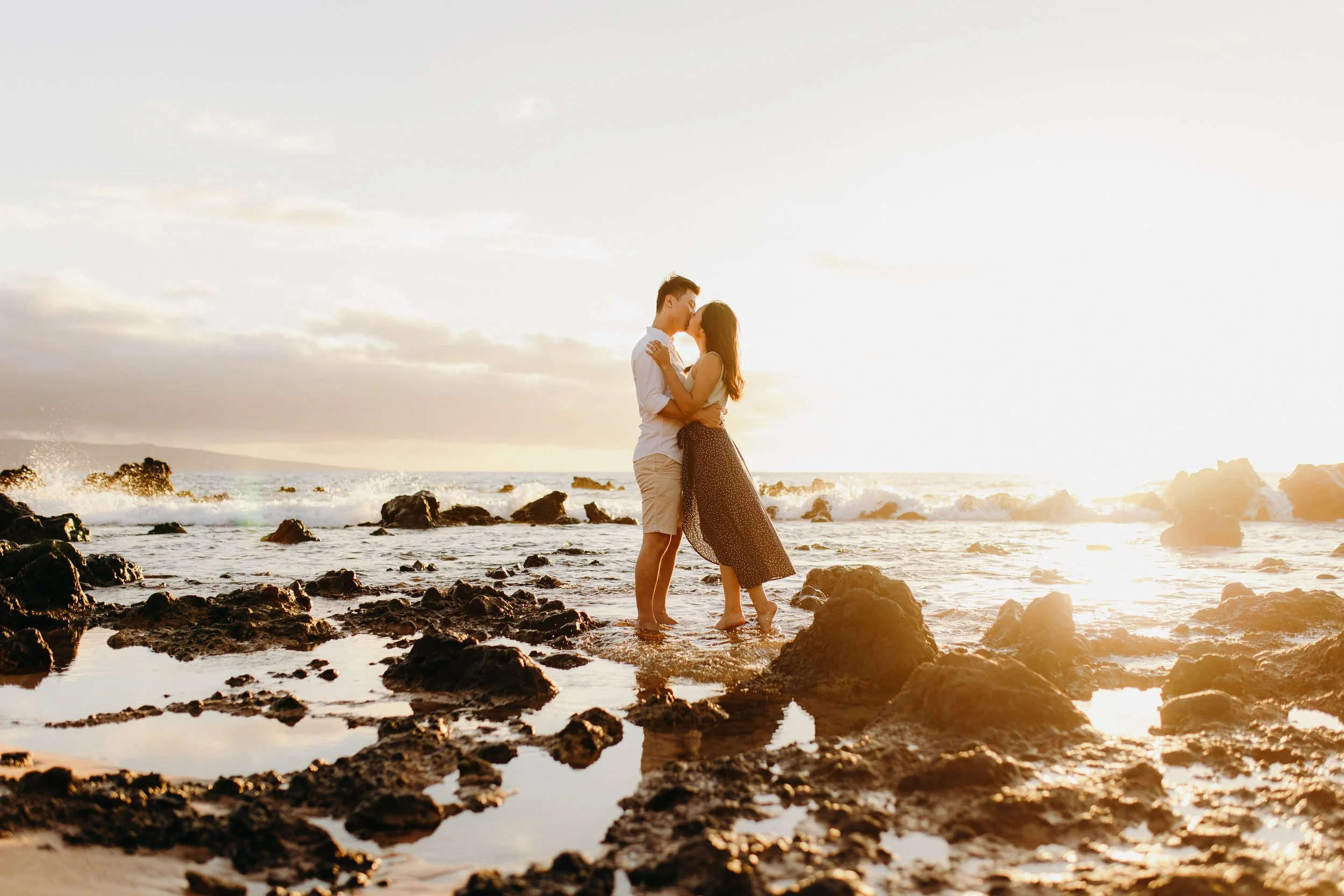 Maui beach portrait of a couple kissing among volcanic rocks at sunset, with ocean waves and golden evening light