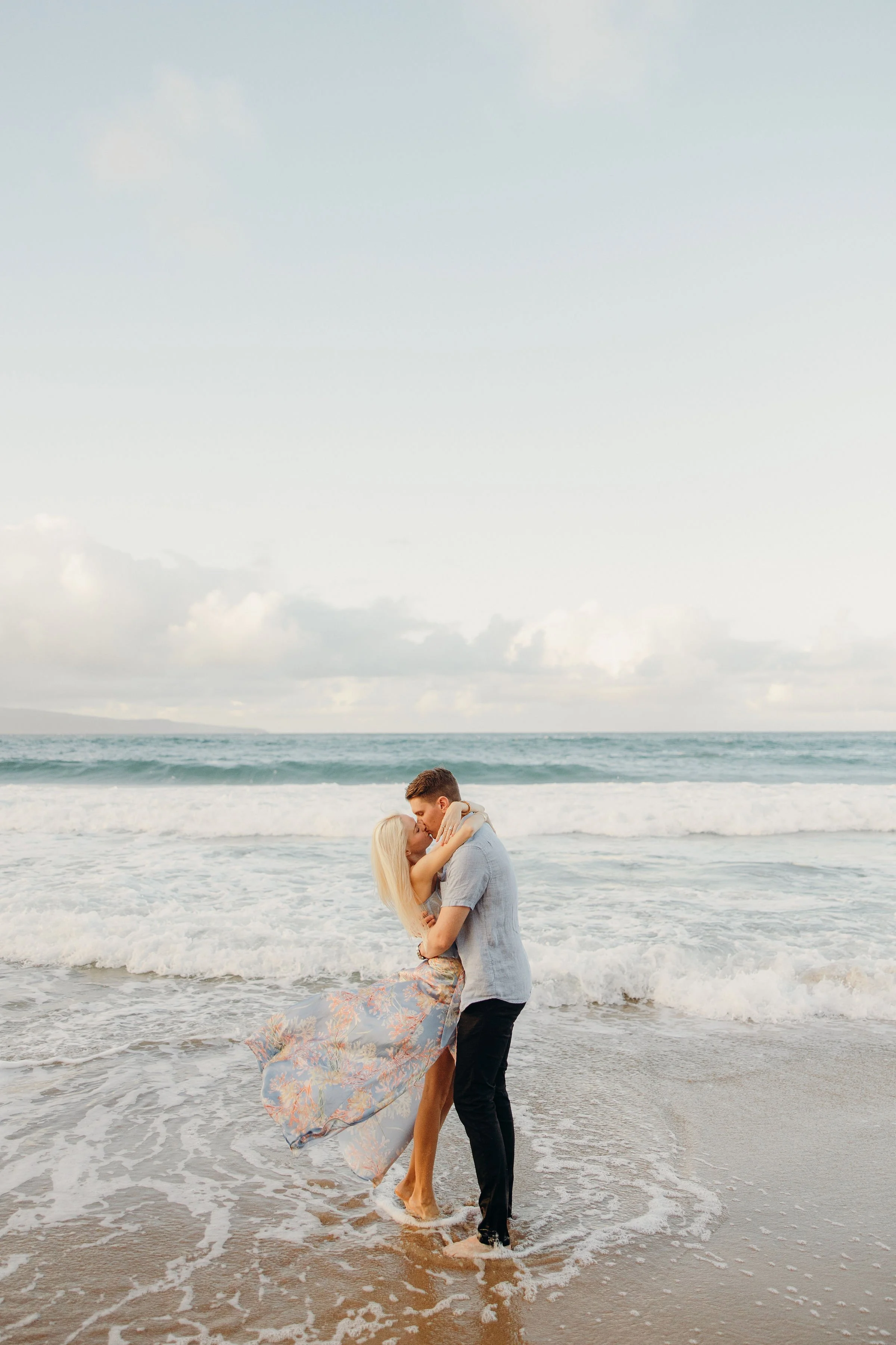 Couple embraces in the ocean surf during a romantic beach portrait session, with waves rolling in behind them.