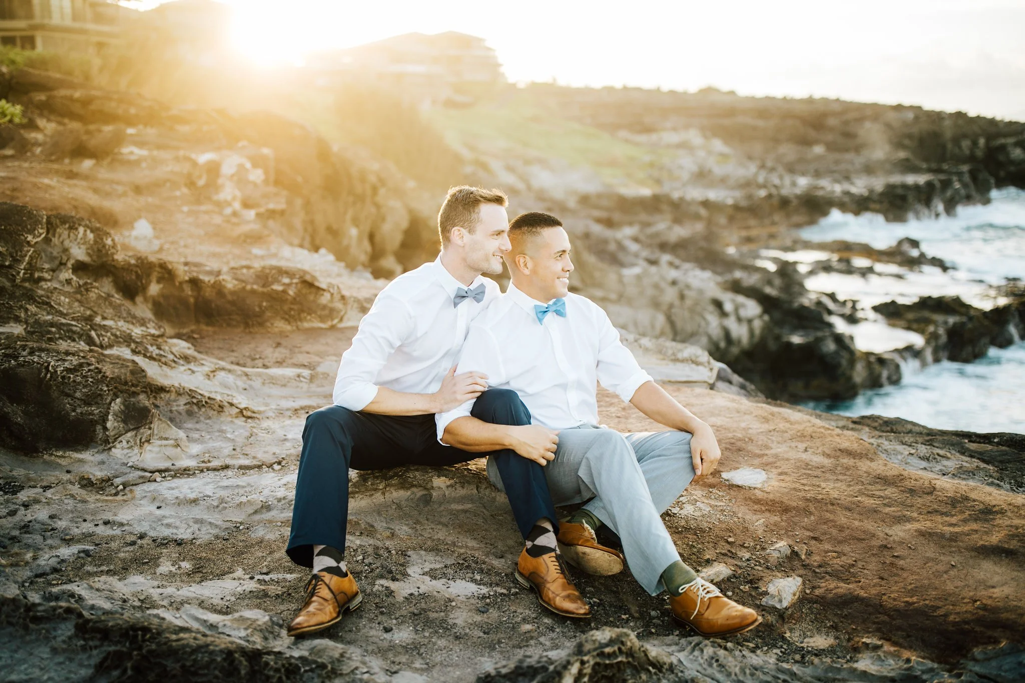 Two grooms in bow ties sit together on rocky shoreline cliffs at sunset during a Maui wedding portrait session.
