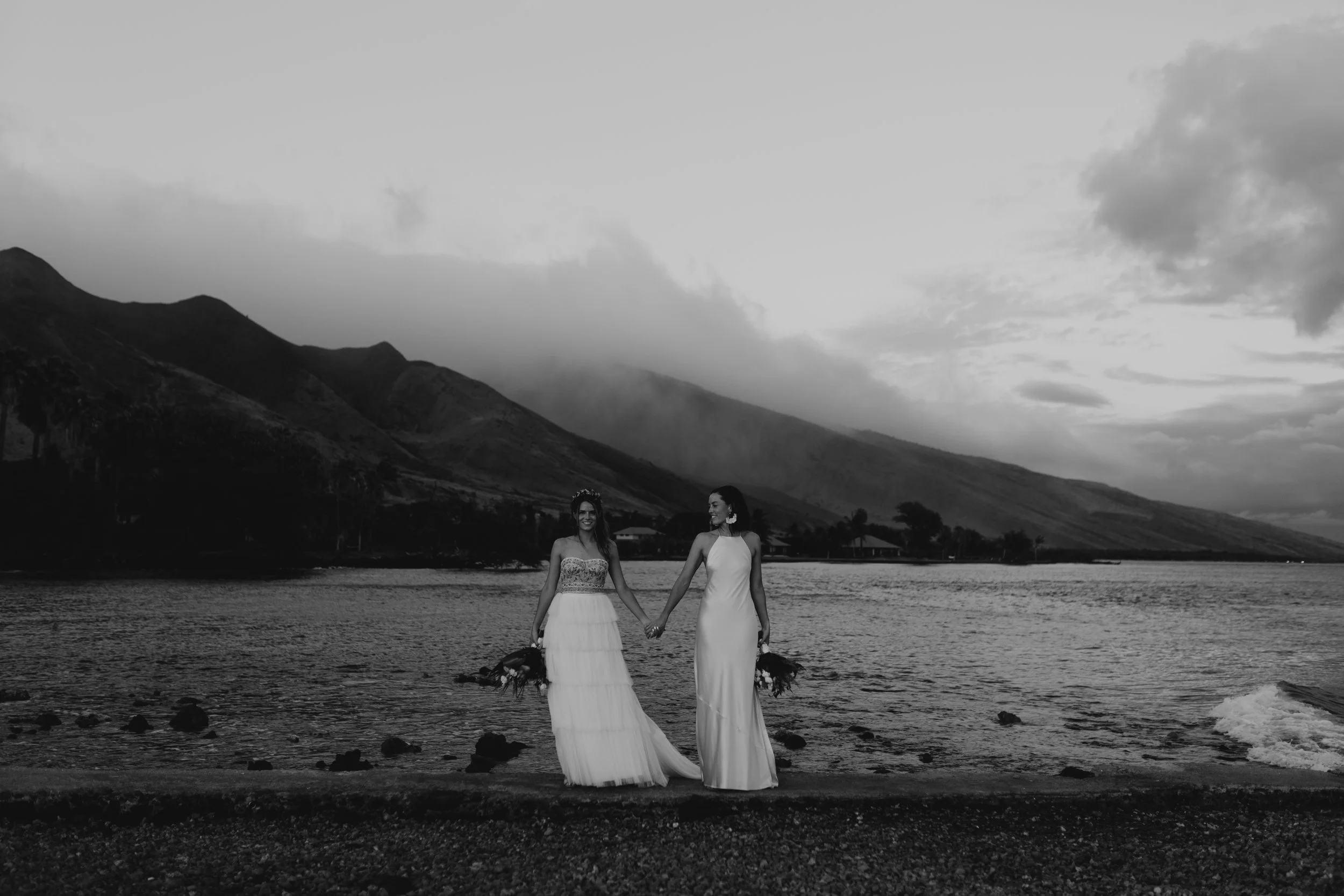 Two brides hold hands on a beach beneath dramatic mountains during a Maui wedding portrait session.