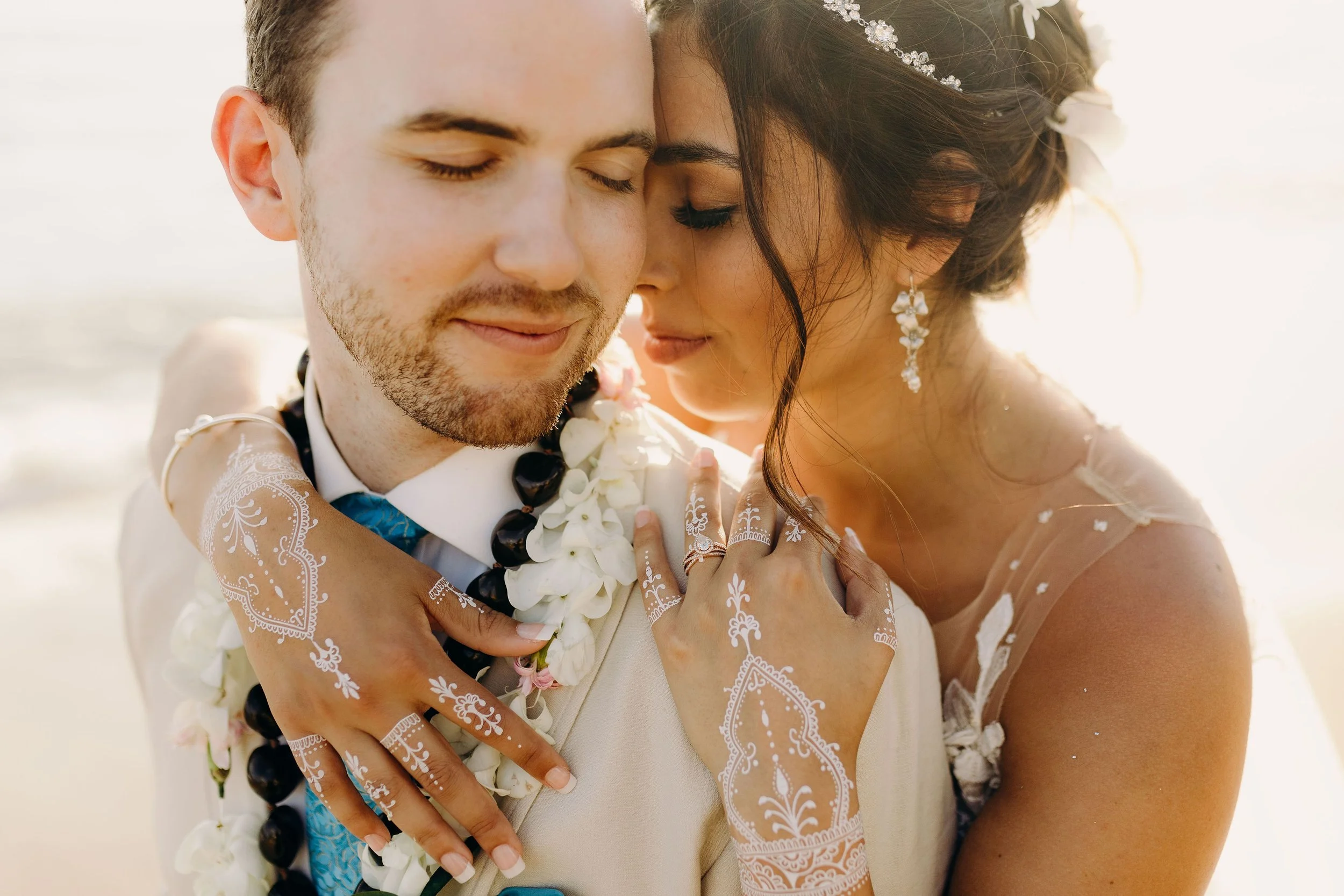 Maui wedding portrait of a bride embracing the groom from behind, showing her lace sleeves, floral lei, earrings, and henna-style hand details