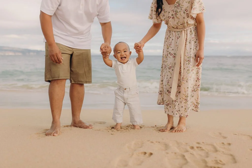 Smiling toddler takes steps on a Maui beach while holding both parents’ hands, with the ocean in the background.