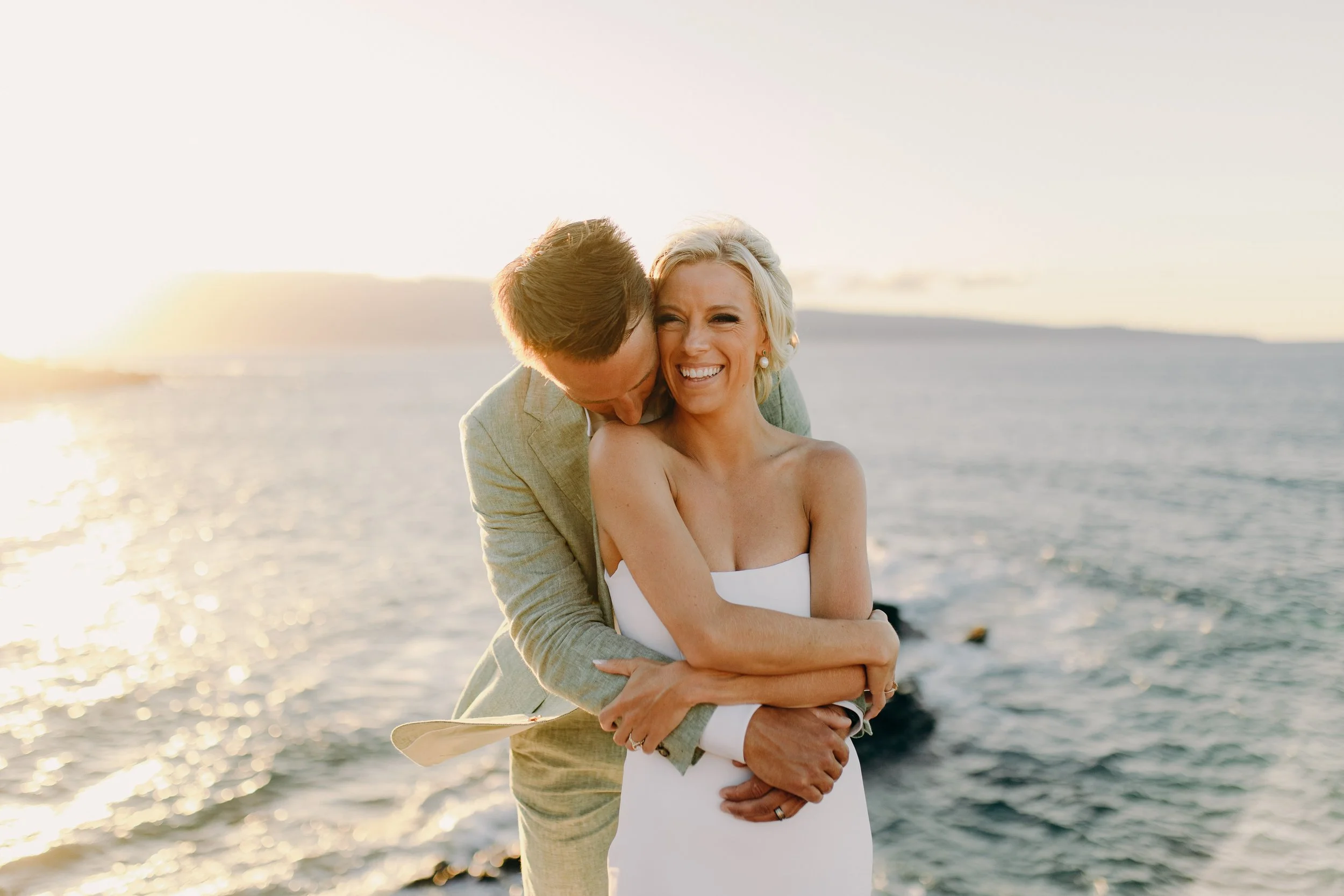 Maui wedding couple embracing beside the ocean, with the groom standing behind the bride in a white strapless gown
