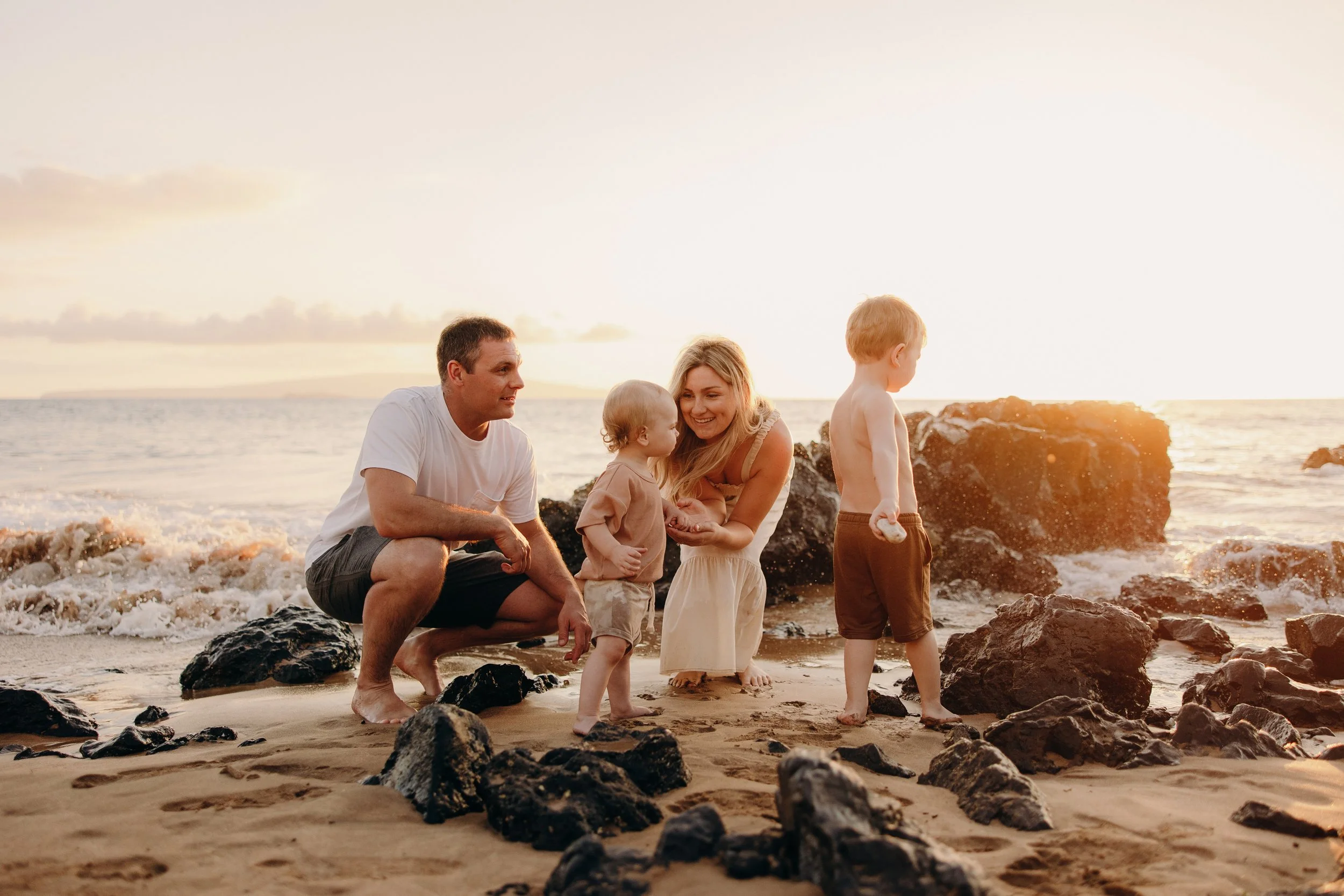 Family of four gathers among dark lava rocks on a Maui beach at sunset, with parents crouching beside their young children near the ocean.