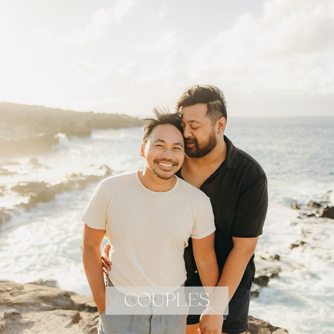 Two men pose on a rocky Hawaii shoreline at sunset, with one partner standing behind the other and kissing his cheek as ocean waves break in the background.