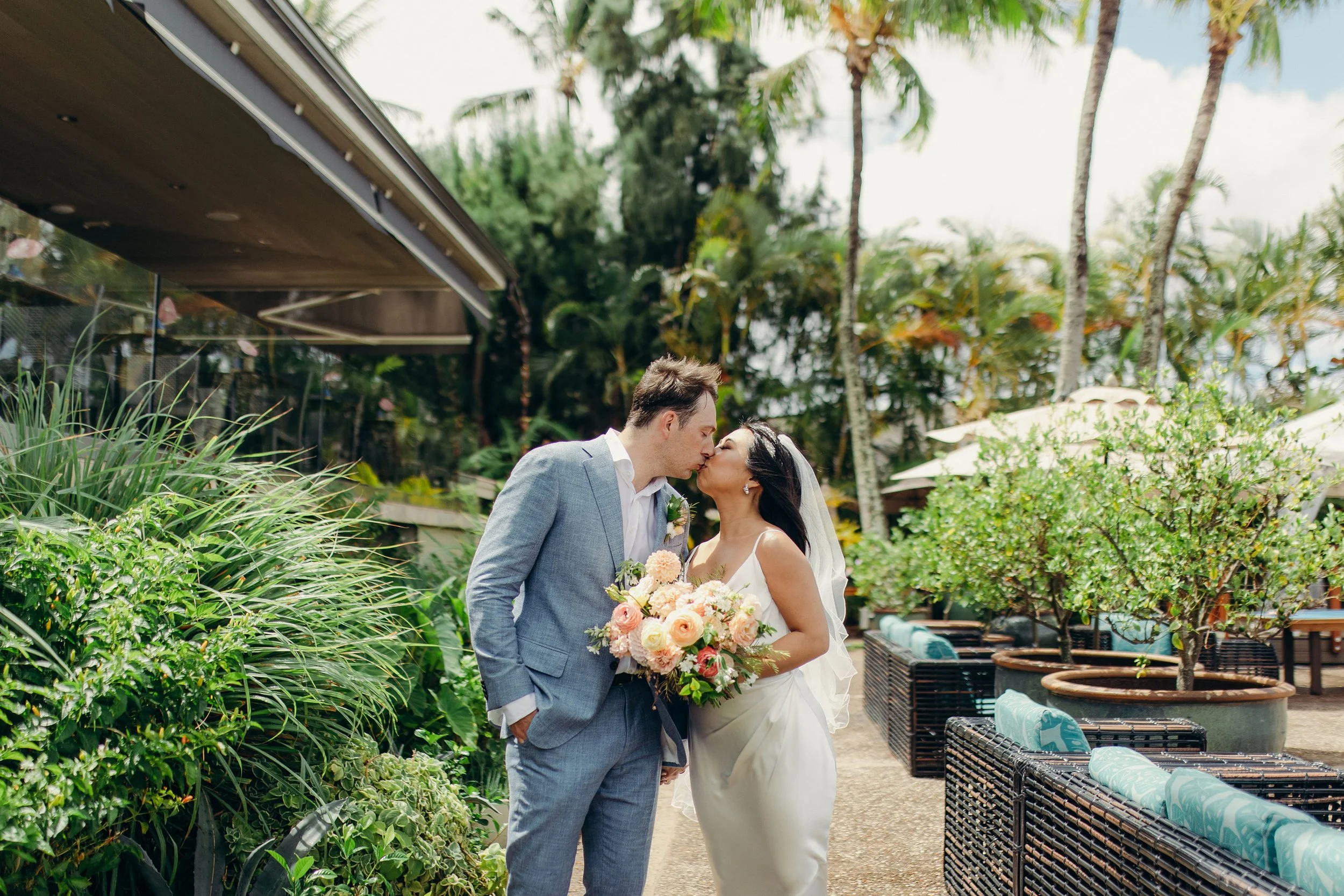 Maui wedding photography of newlyweds kissing in a tropical garden, with the bride wearing a white gown and veil and carrying a peach and cream flower bouquet