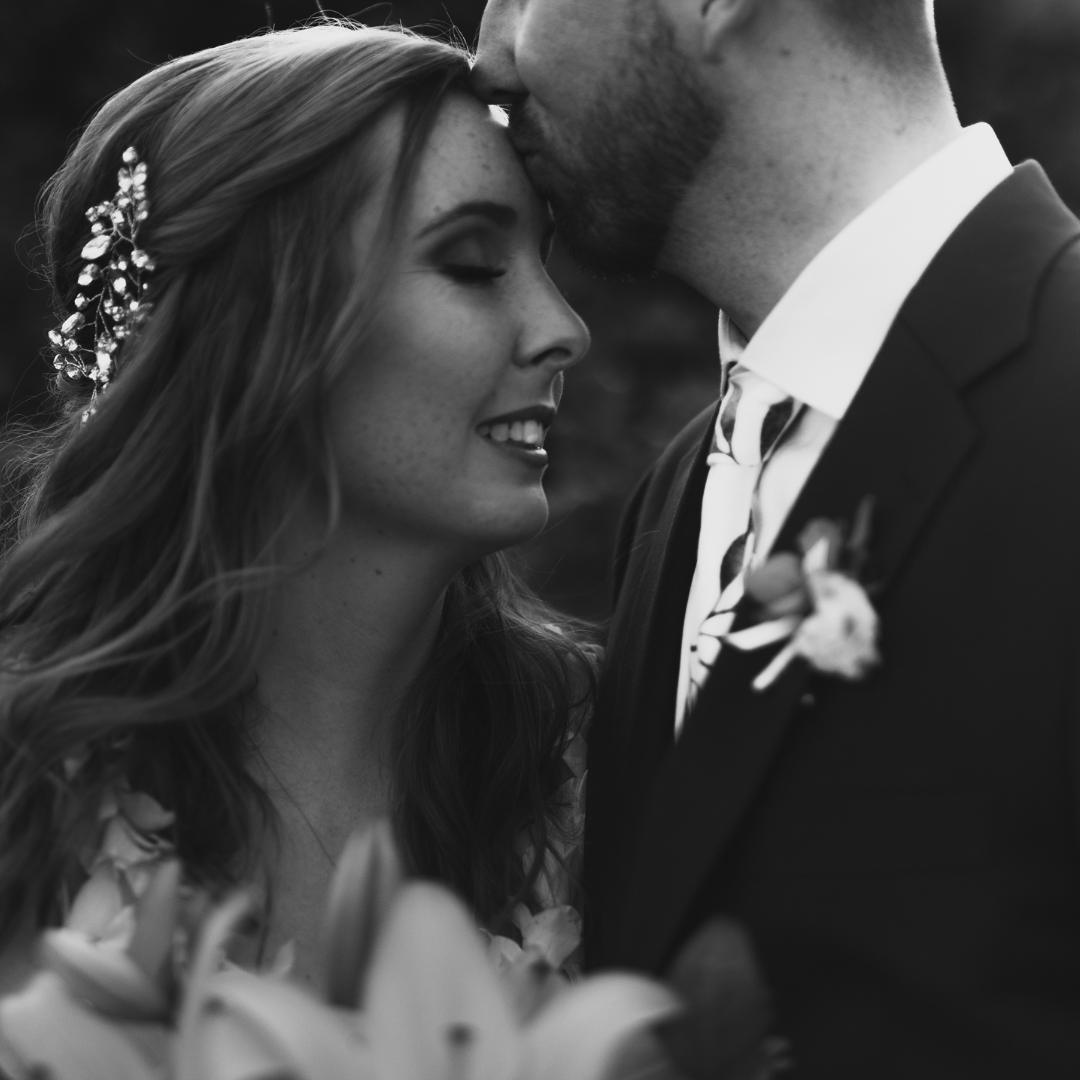 Black-and-white Maui wedding photograph of a bride smiling as the groom kisses her forehead, with a jeweled hairpiece and boutonniere