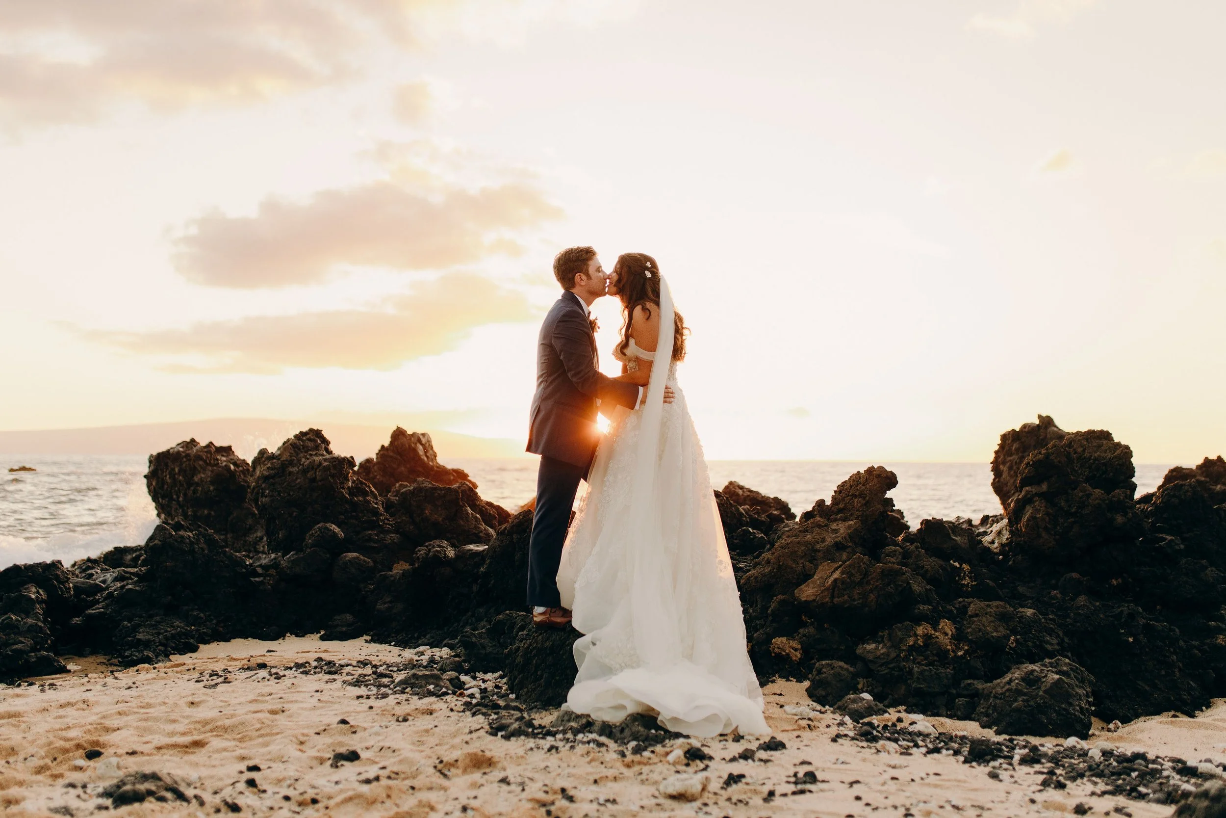 Maui wedding couple kissing on a rocky beach at sunset, with the bride wearing a flowing white gown and the groom in a dark suit