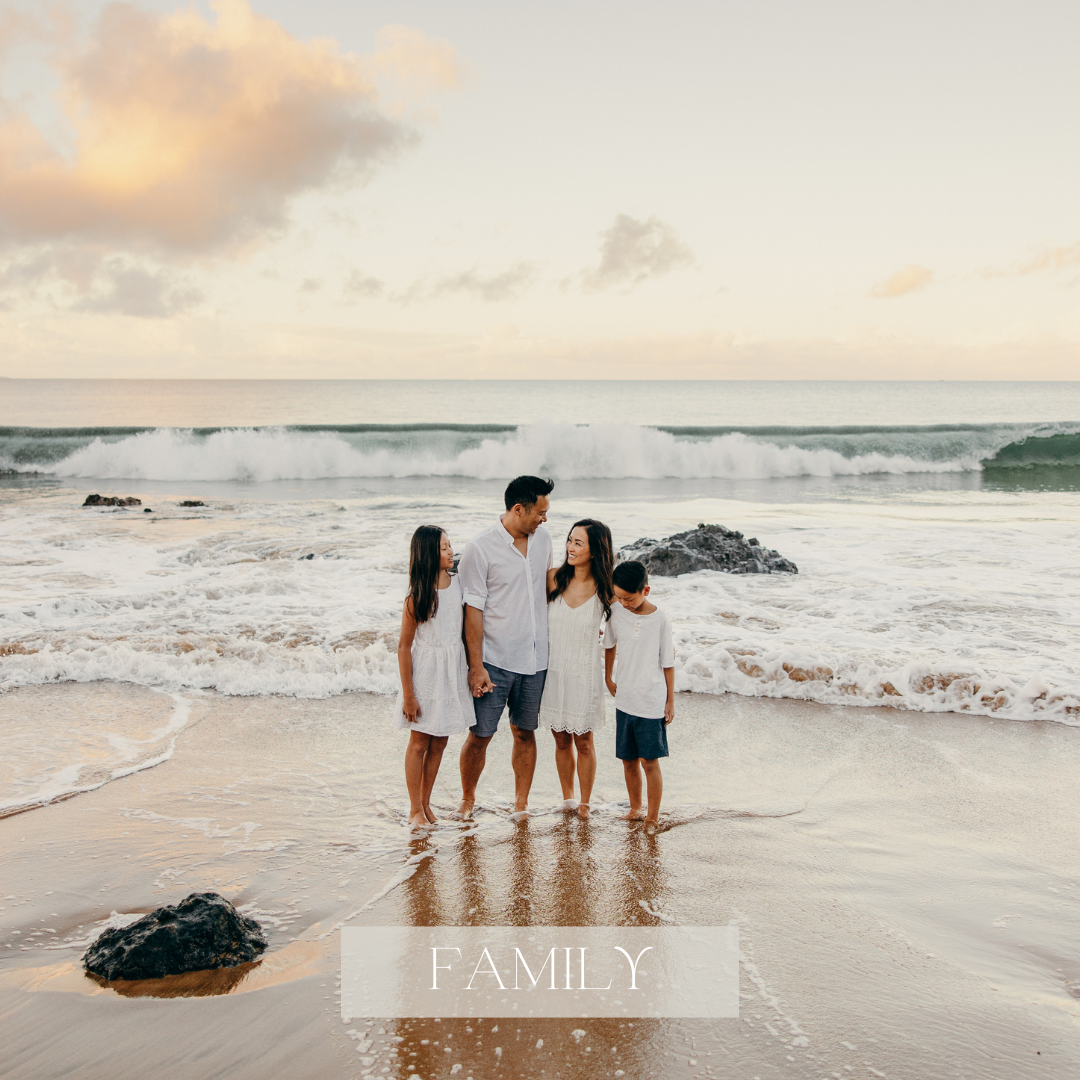 Family of four stands together on a Maui beach with their feet in the ocean, surrounded by waves and rocky shoreline.