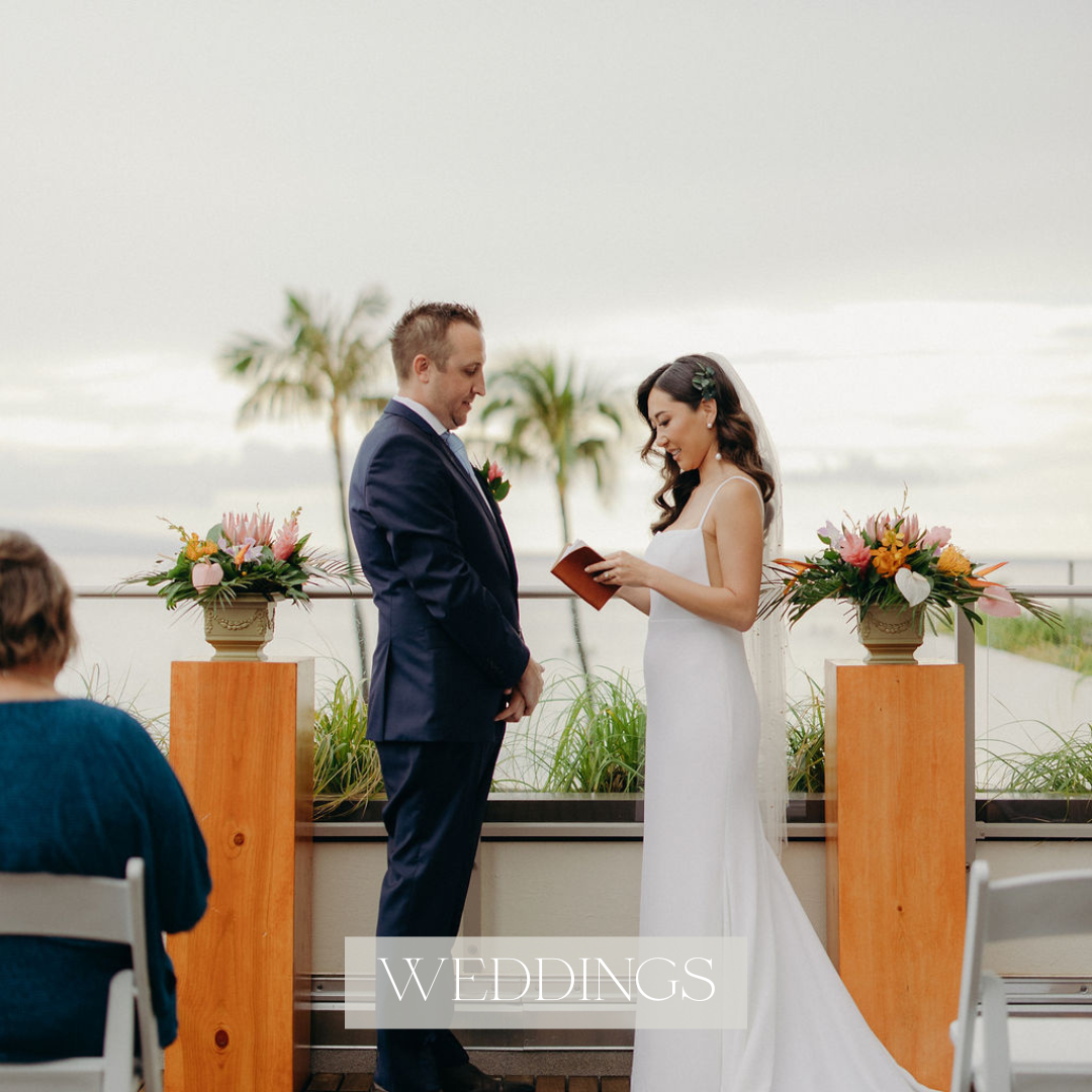 Bride in a white gown holds a colorful tropical bouquet while facing the groom in a navy suit during an outdoor Hawaii wedding ceremony, with palm trees and the ocean behind them.