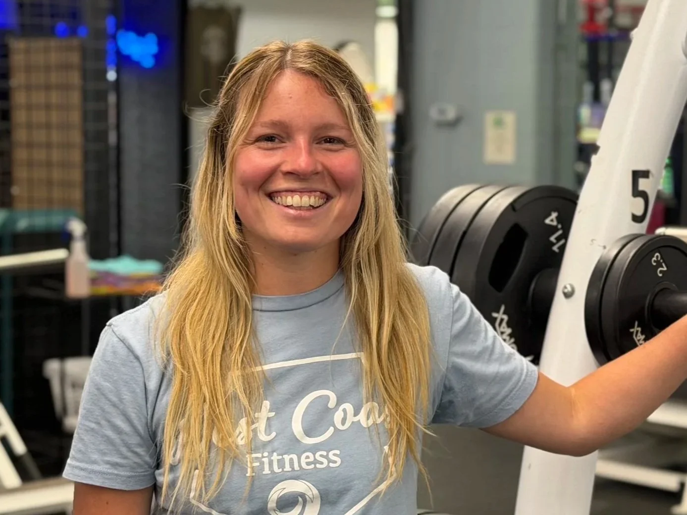 Smiling woman in a blue West Coast Fitness T-shirt standing in a gym with exercise equipment in the background.