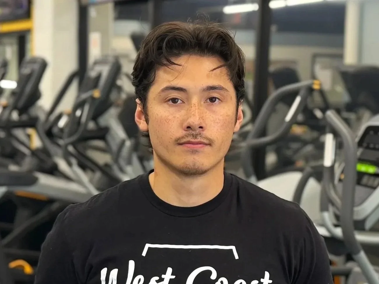 A young man with dark, curly hair and light brown skin standing indoors with cardio equipment