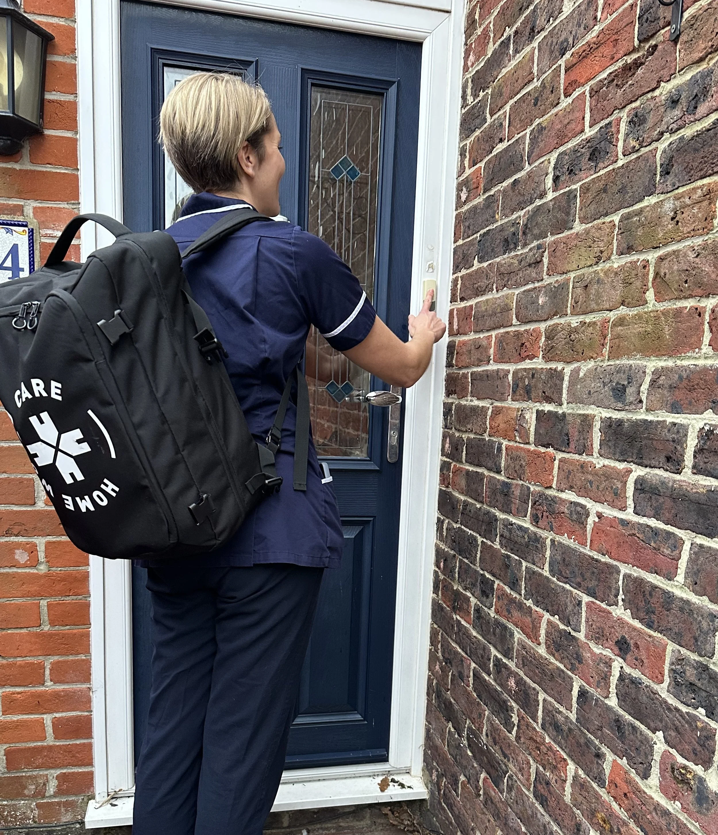 A woman in uniform with a backpack ringing a doorbell at a house with a brick wall and a blue front door.