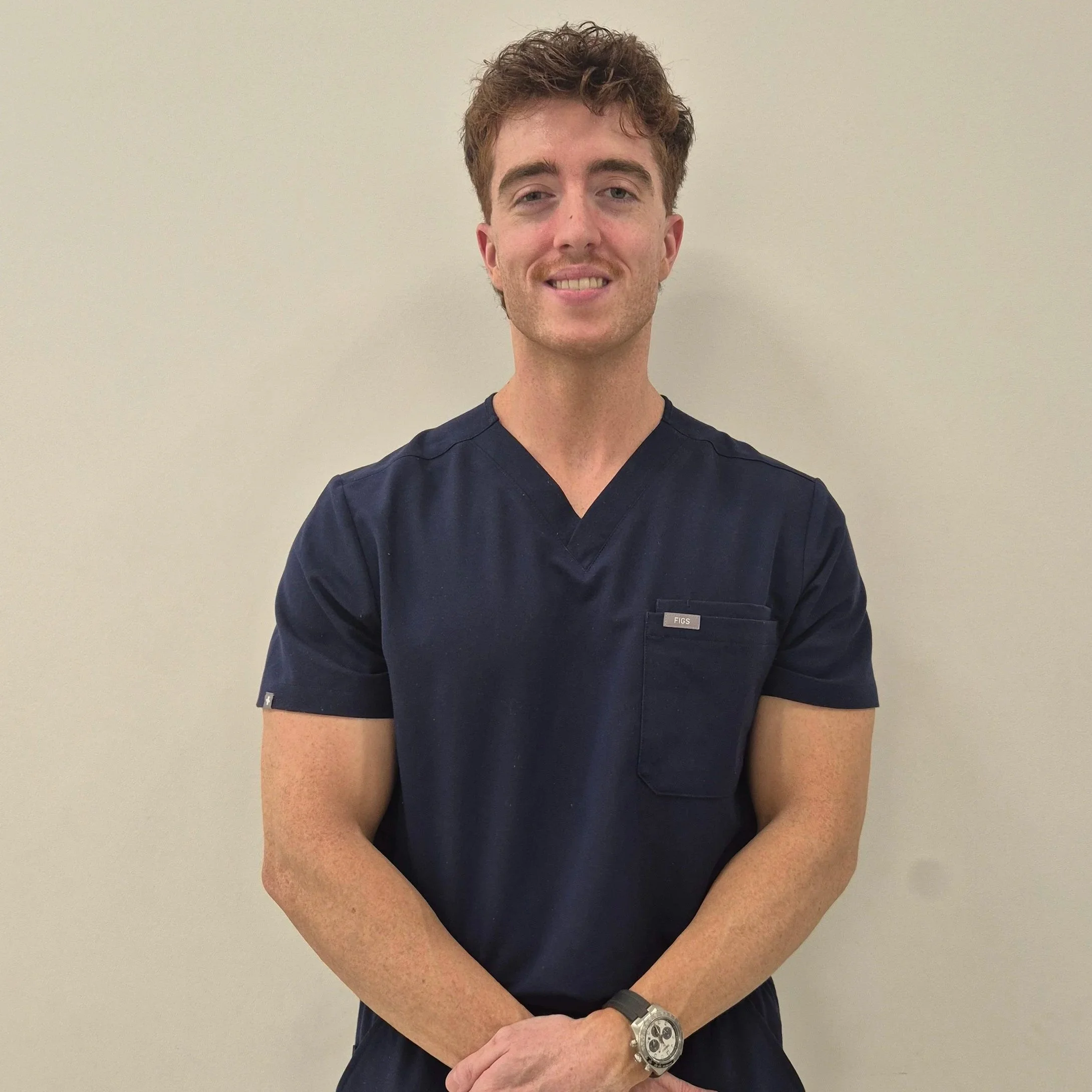 A young man with curly brown hair and light skin stands against a plain light-colored wall. He is smiling slightly, showing his teeth. He wears a dark scrub top with a small logo on the pocket and a large silver watch on his left wrist. His arms are crossed, and he looks directly at the camera.