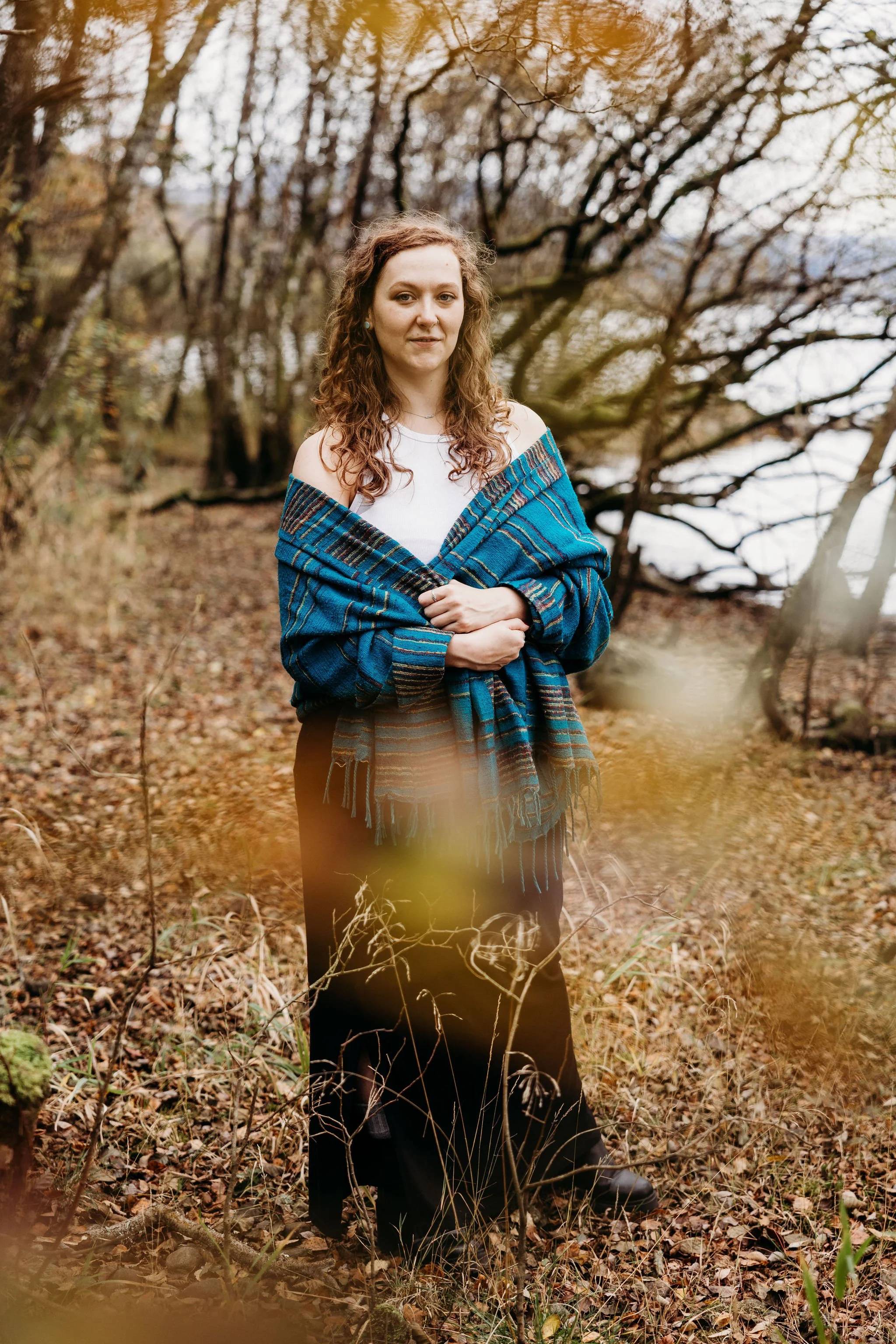 A young woman standing in an autumnal forest, wearing black pants, a white top, and a blue plaid shawl, looking at the camera with her arms crossed over her chest.