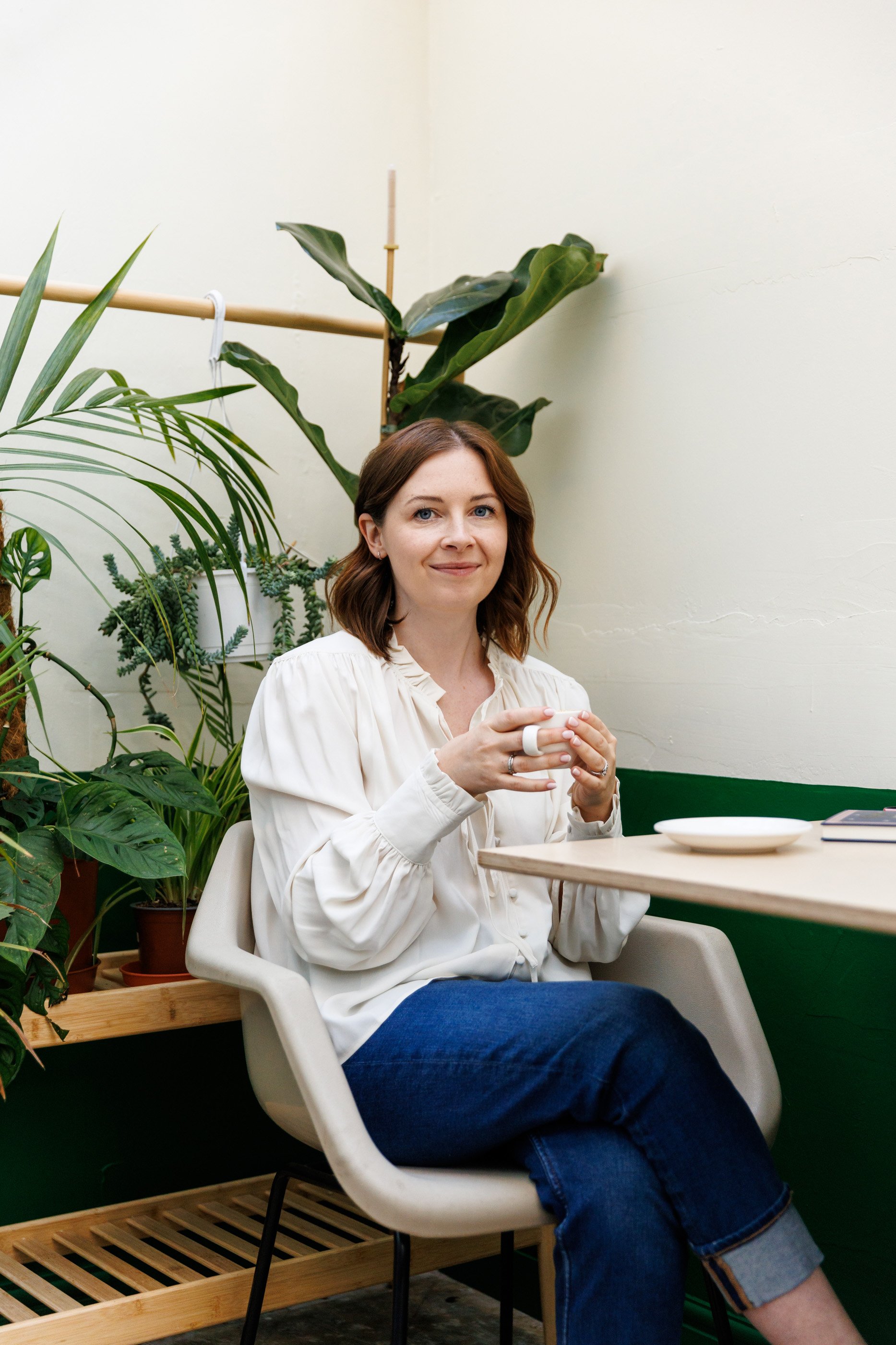 Woman in a white blouse sitting in a chair and holding a cup, surrounded by indoor plants.