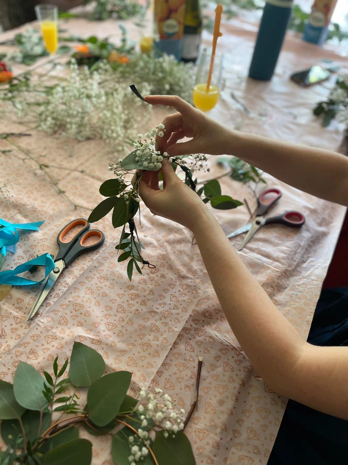 Person arranging white baby's breath flowers with green leaves on a table covered with patterned paper, surrounded by scissors and ribbons, with drinks and other floral arrangements in the background.