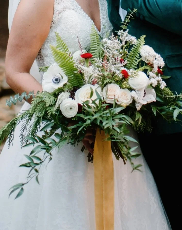 christmas - winter-themed bridal bouquet featuring soft white blooms mixed with lush evergreens and fern fronds, white anemones with dark centers, white ranunculus, and light roses, eucalyptus branches, small red flower or berry accents