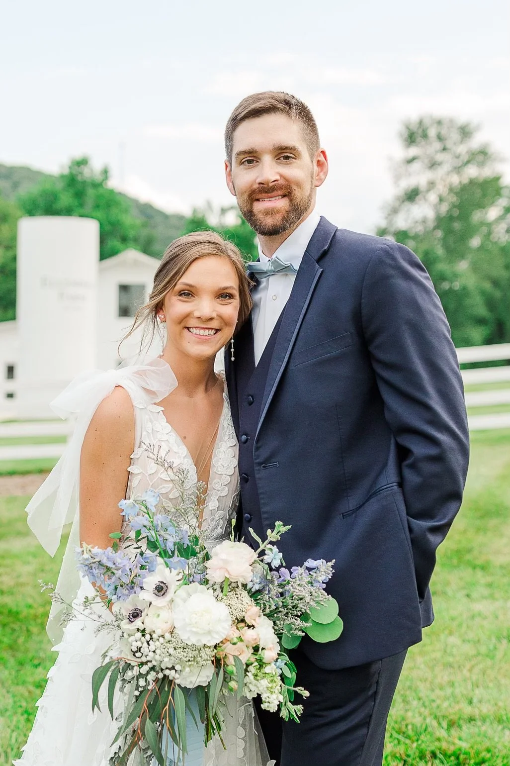 Bride and groom in front of an old farm cilo. The bride is holding a bouquet of flowers blue tones of delphinium and tweedia flowers 