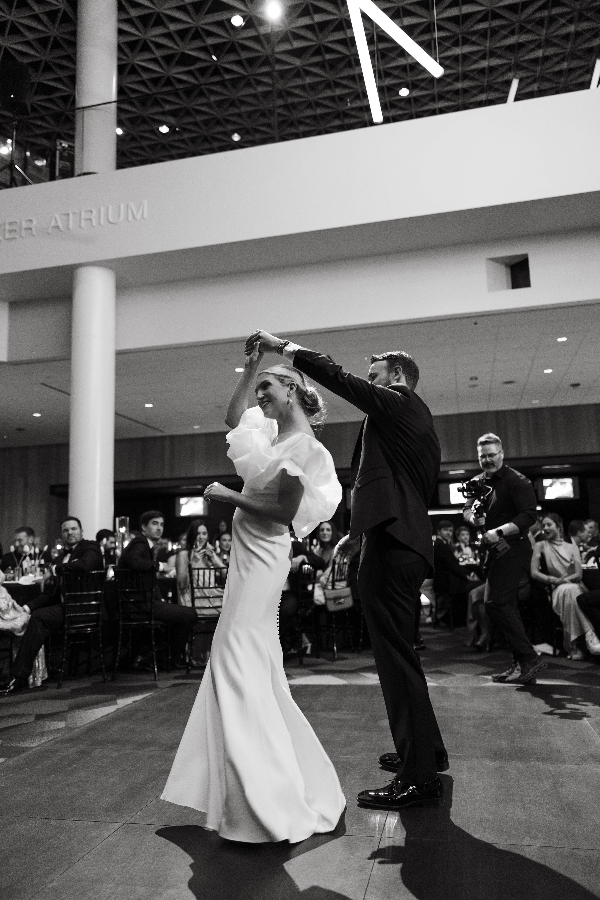 Black and white photo of a bride and groom dancing at their wedding reception with guests seated around them, in an indoor venue.