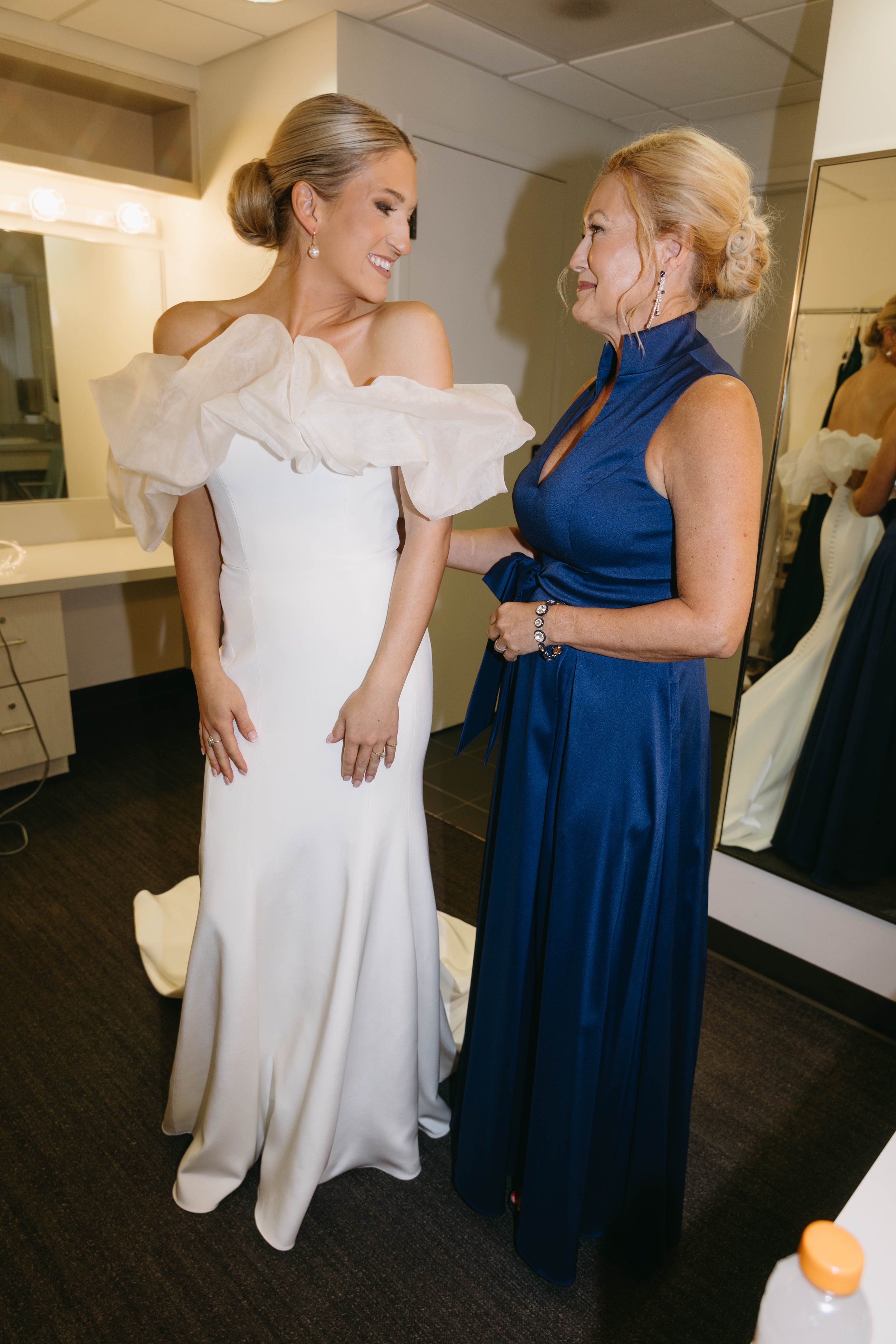 Two women in a dressing room; one in a white wedding dress with ruffled off-the-shoulder detail, the other in a blue gown. They are smiling at each other.