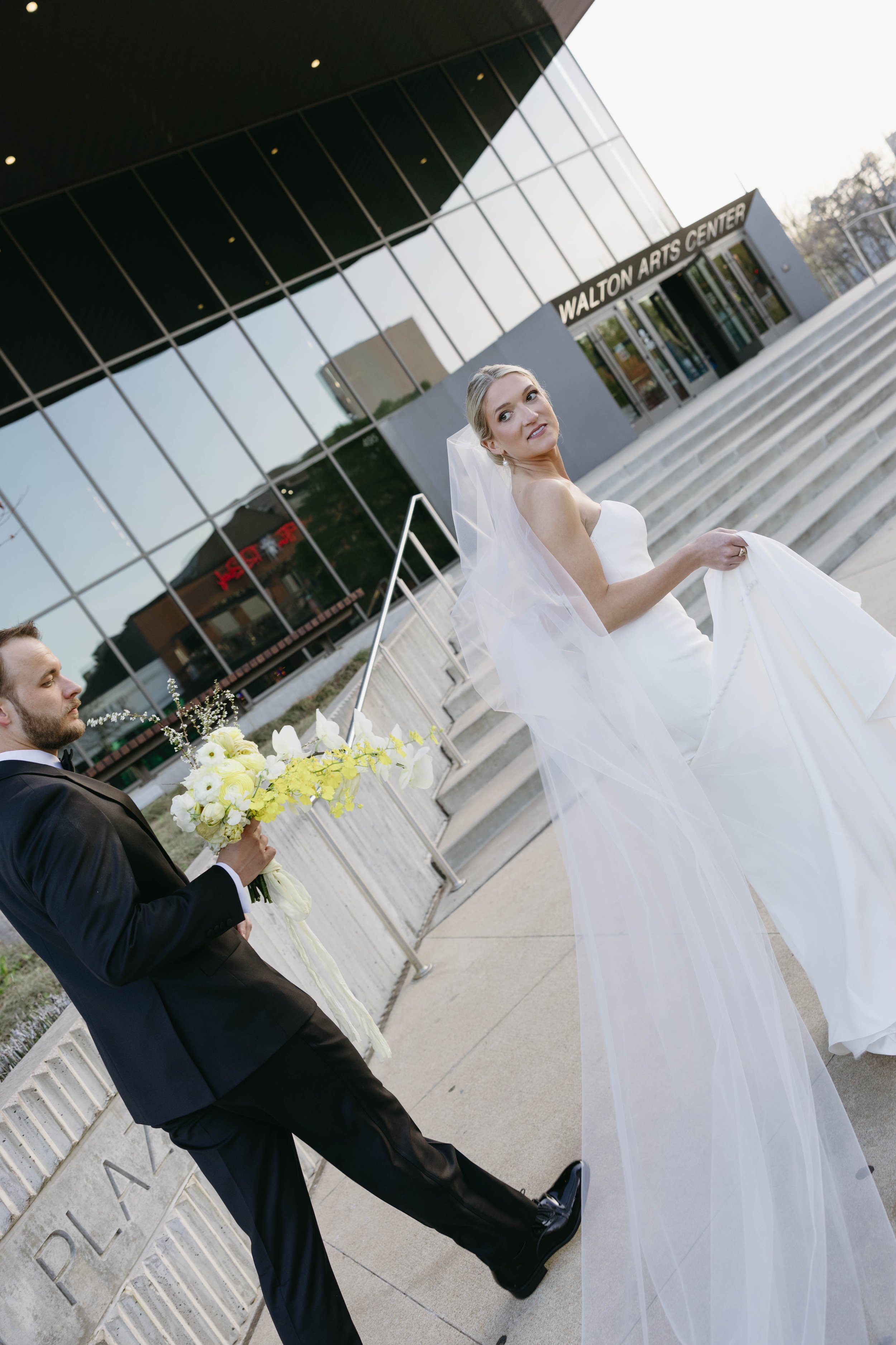 Bride and groom outside Walton Arts Center, bride in strapless gown holding veil, groom in black suit holding bouquet.