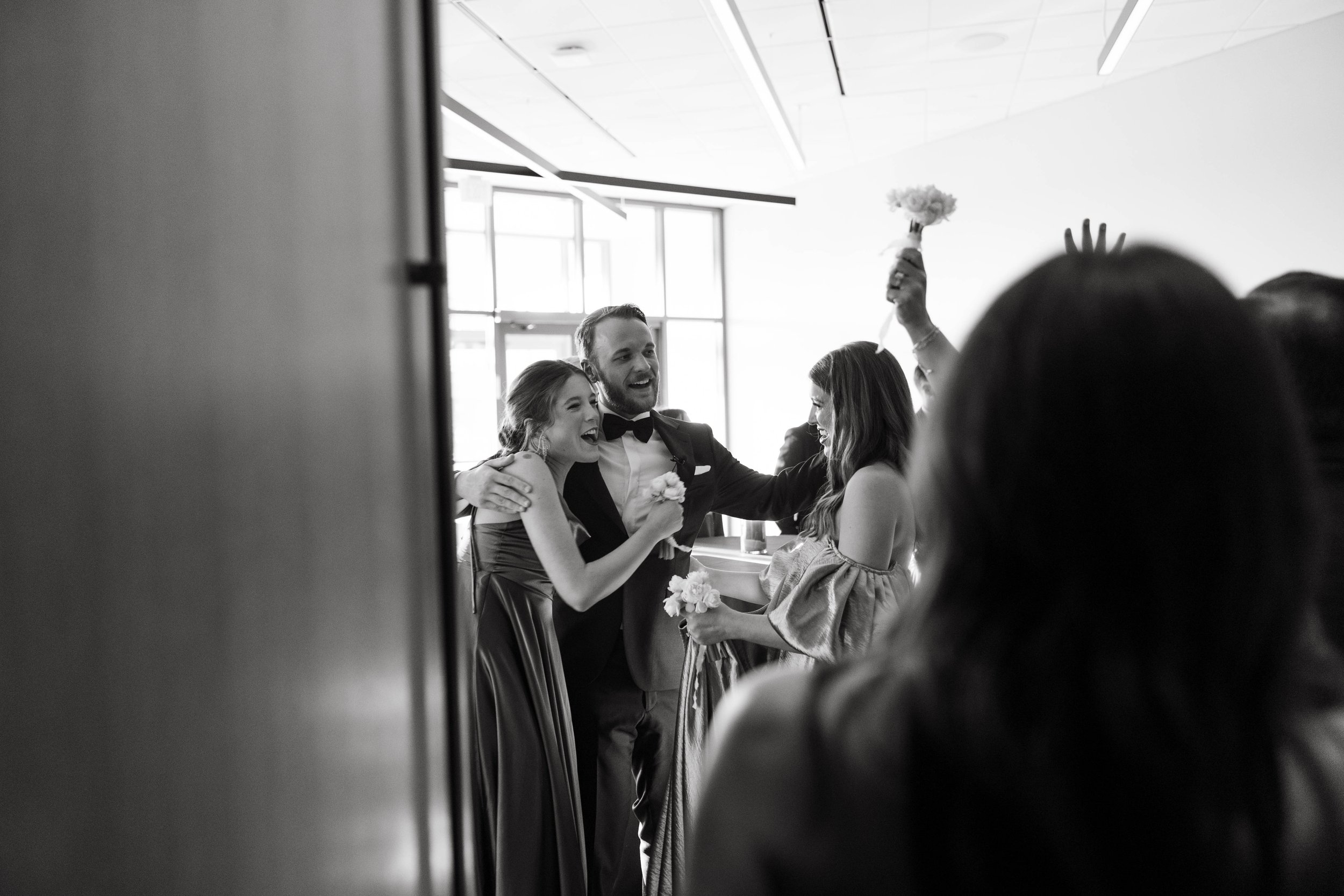 A group of people in formal attire, smiling and celebrating indoors. A man in a suit has his arms around two women in dresses, one of whom is holding a bouquet.