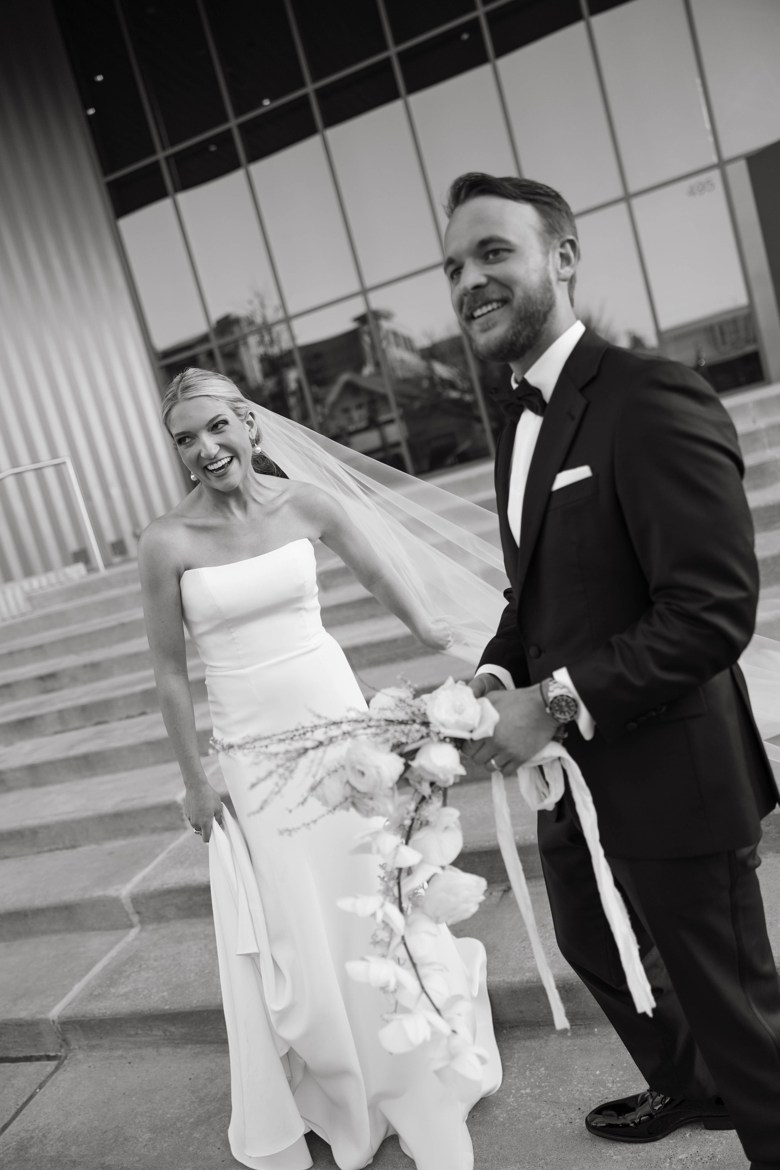 Bride in white gown and veil standing beside groom in tuxedo, holding bouquet, laughing outdoors, black and white photo.