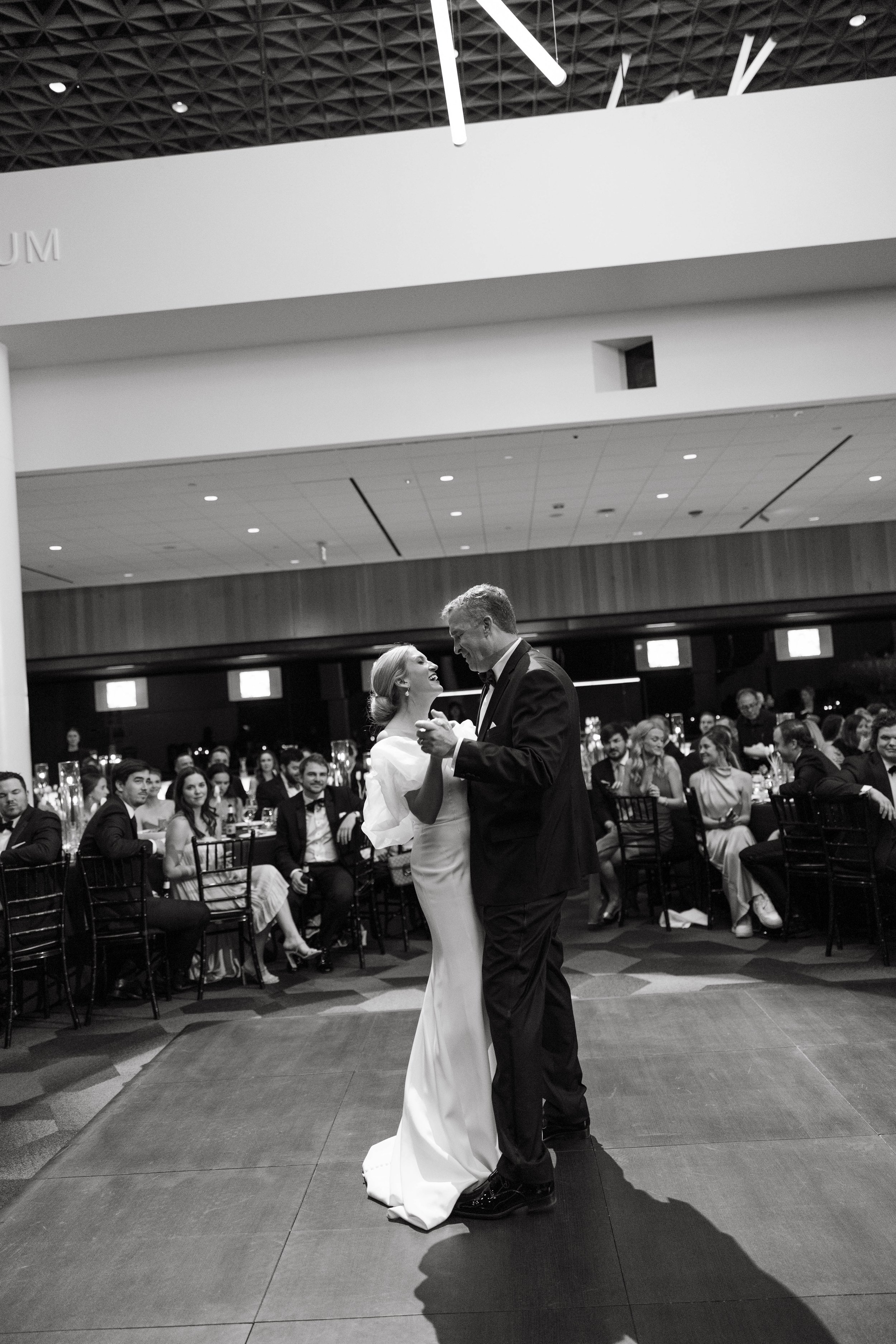 A black and white image of a bride and groom dancing at a wedding reception, surrounded by seated guests. The venue has a modern interior with a high ceiling.