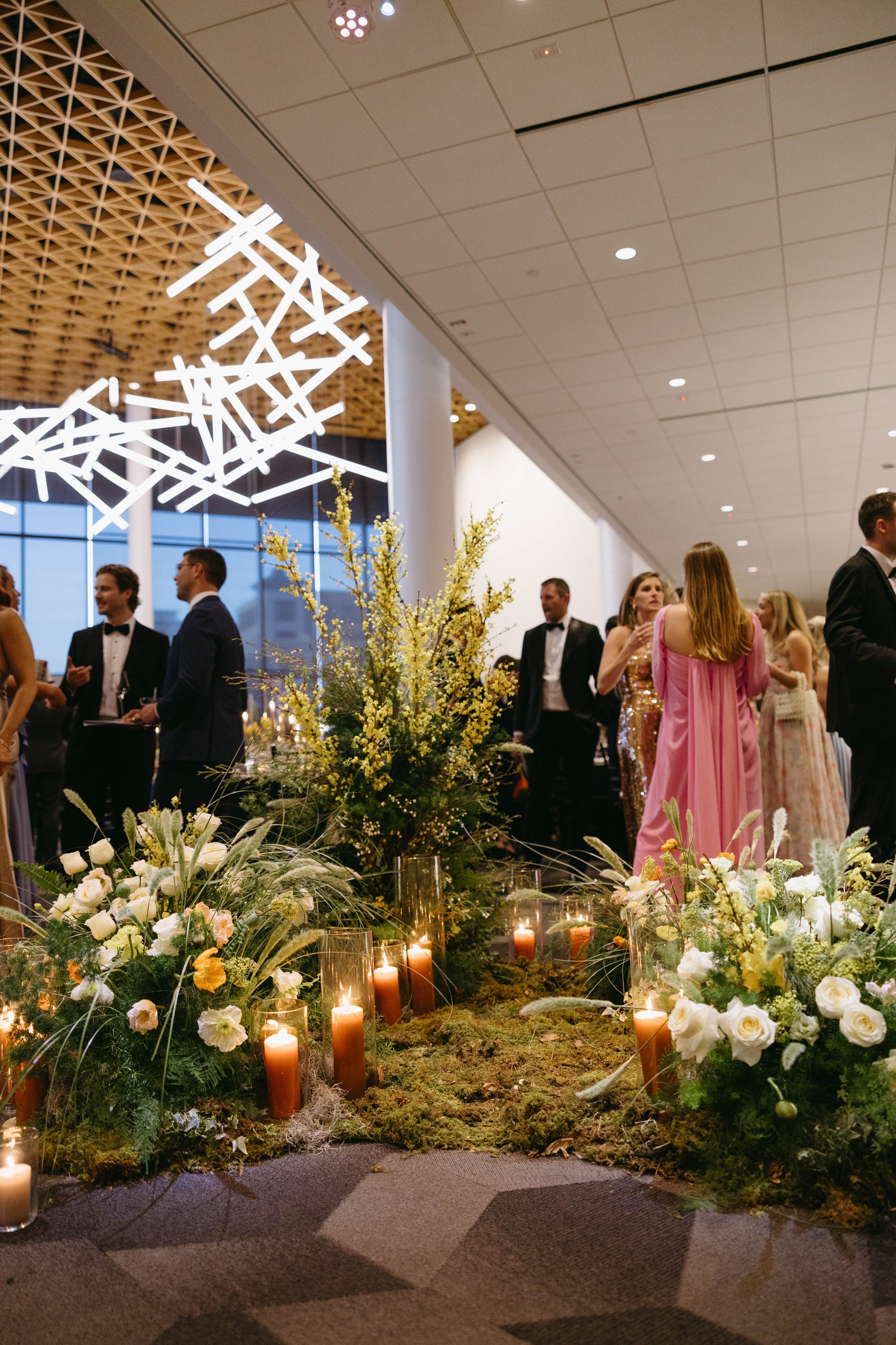 People in formal attire at an indoor event with floral arrangements and candles.