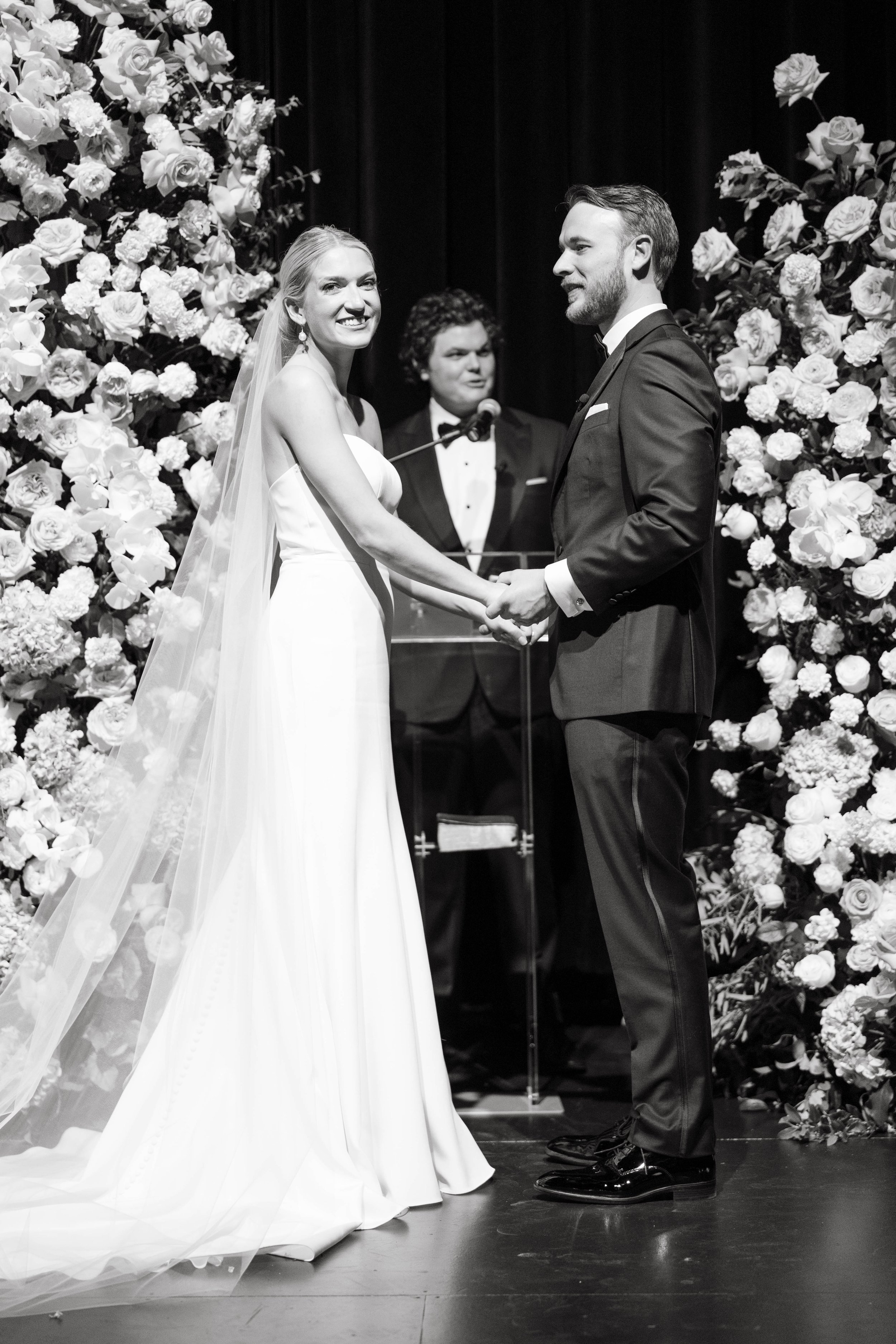 Bride and groom at wedding ceremony, holding hands, with floral arrangements and officiant in the background.