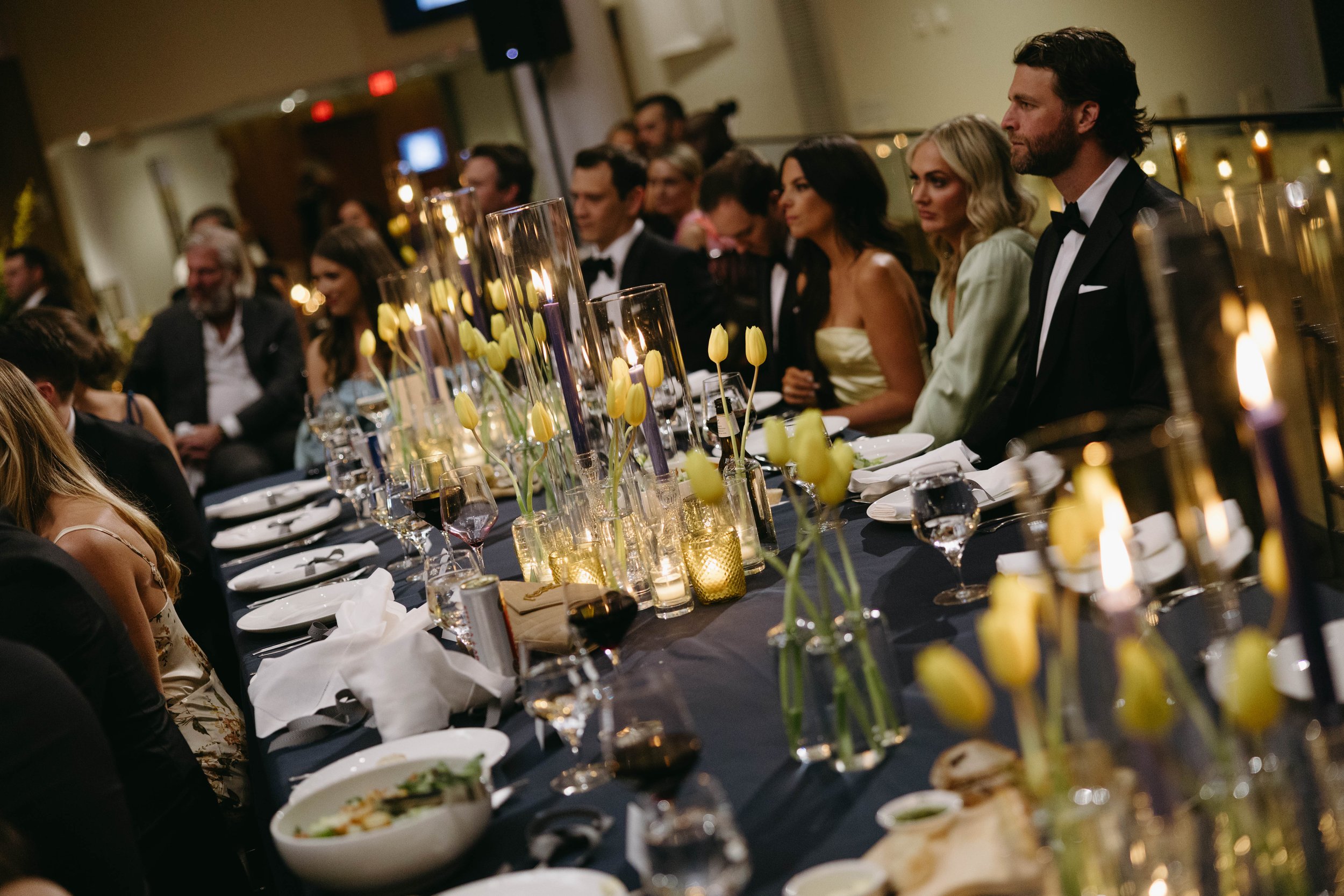 Elegant dinner party scene with guests seated at a long table adorned with yellow tulips, candles, and place settings. Attendees are dressed in formal attire, suggesting a special occasion or event.