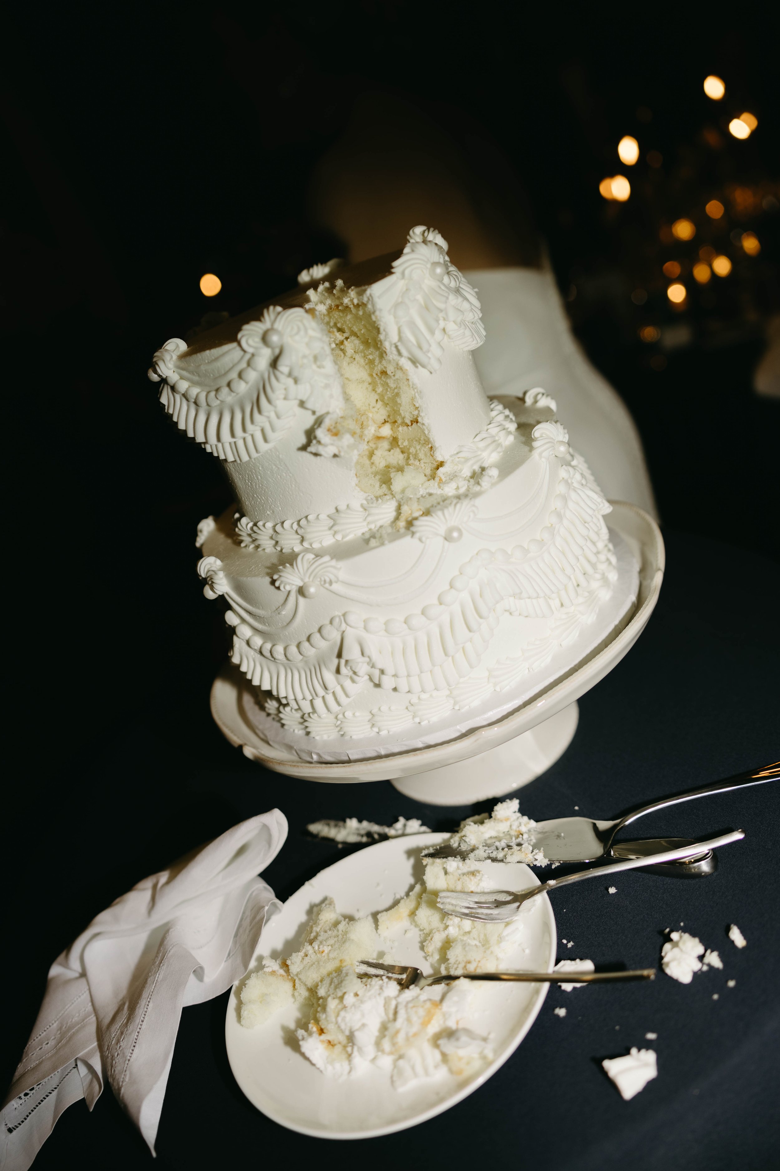 Partially eaten white tiered cake on a stand with pieces and crumbs scattered on a plate and table, accompanied by forks and a white cloth napkin.