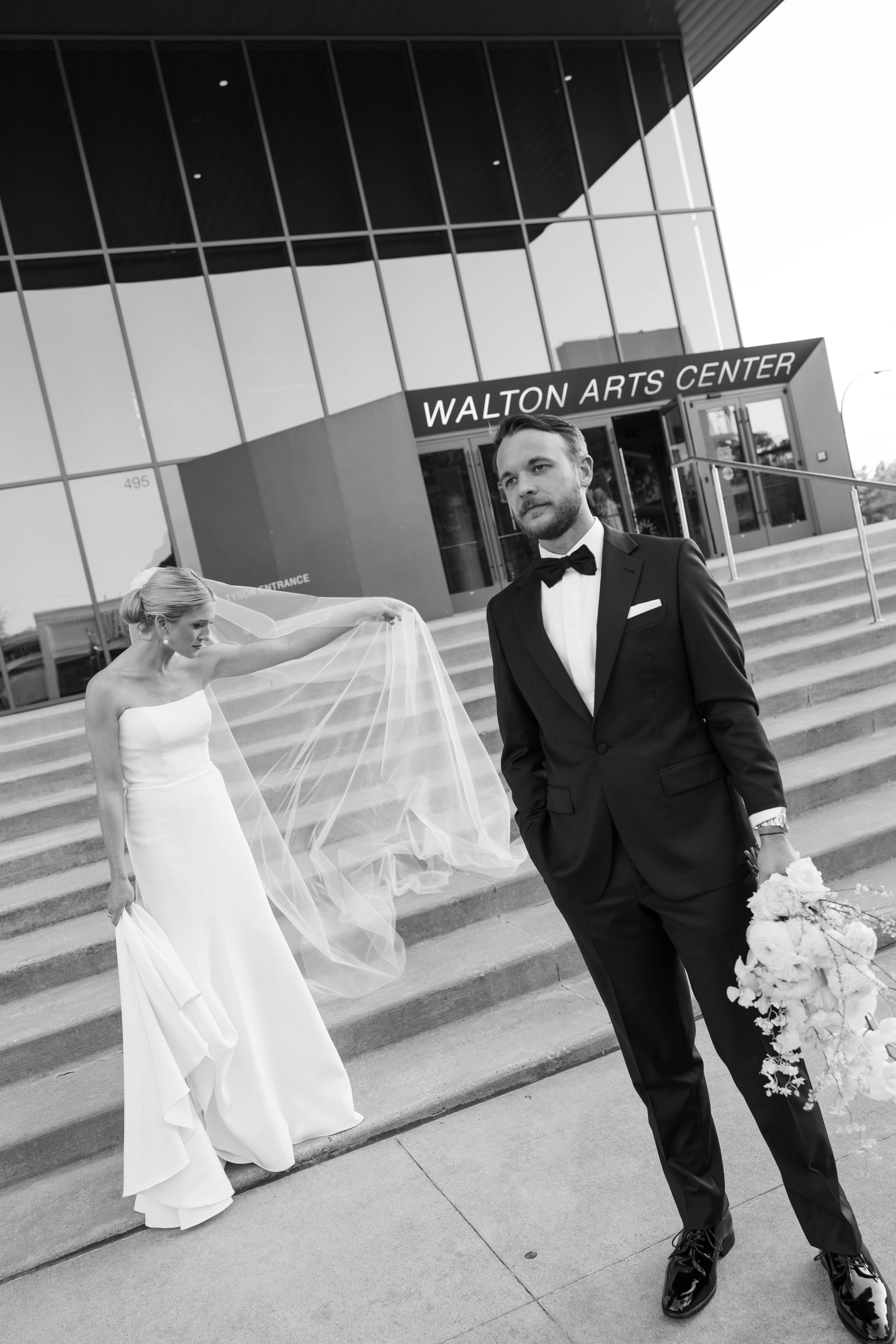 Bride and groom posing outside Walton Arts Center steps, bride holding veil, groom holding bouquet, black and white photo.