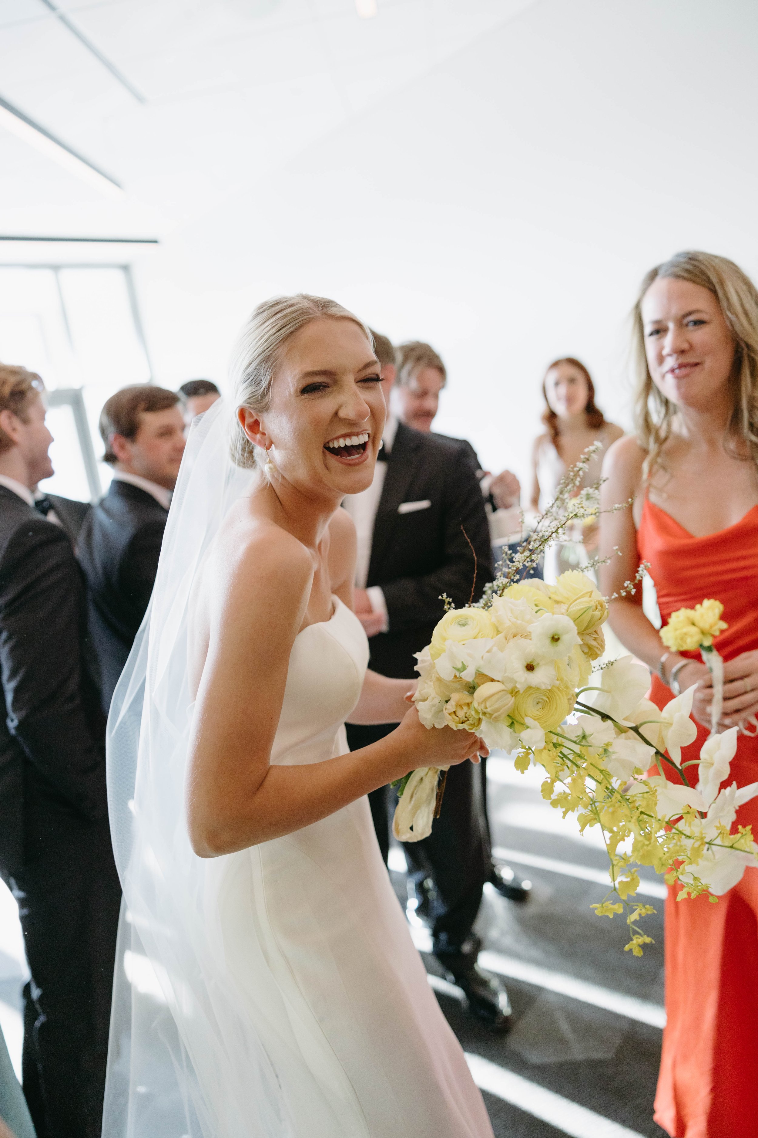 Bride in white dress laughing and holding a bouquet with people in background at a wedding.