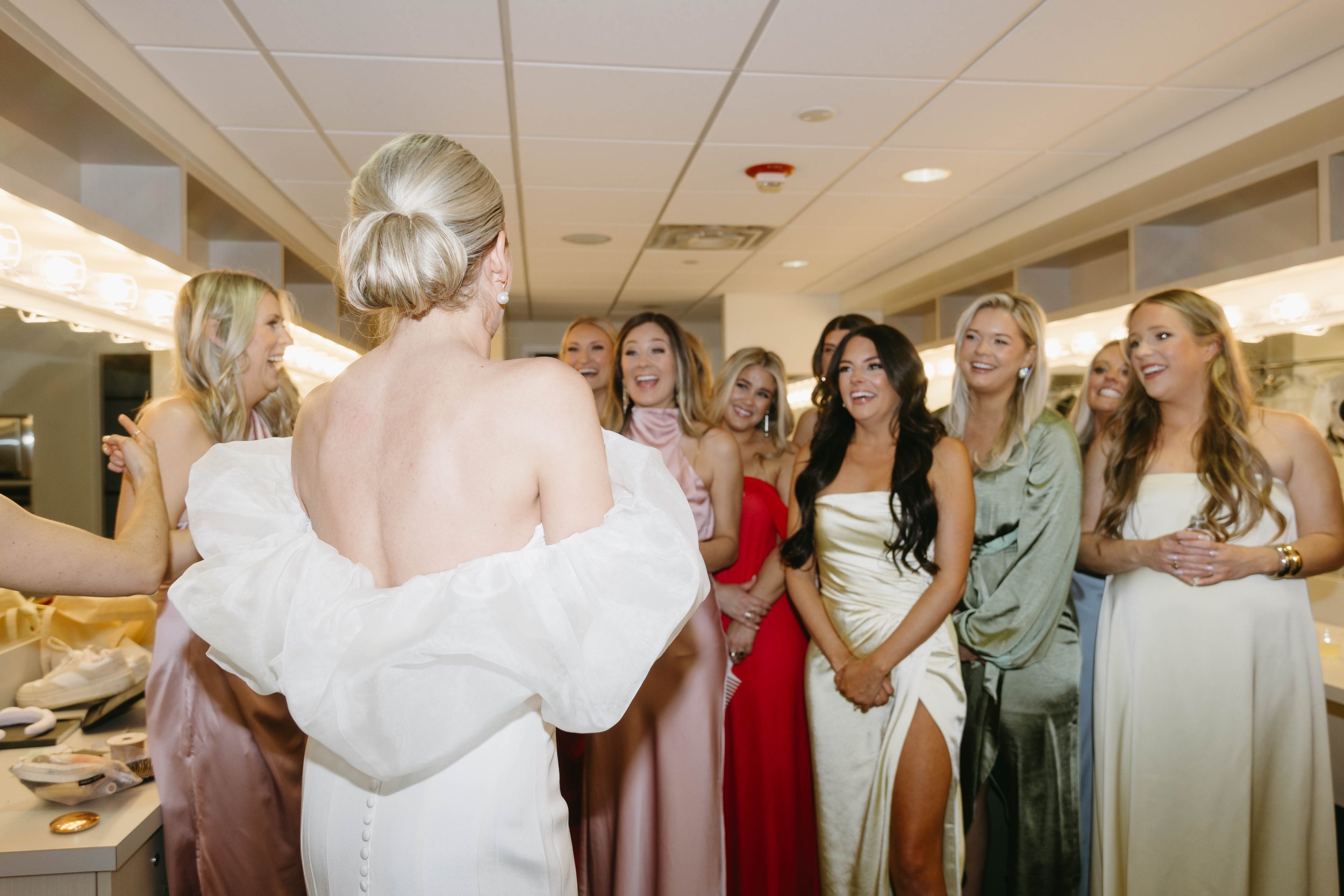 A group of women in formal dresses gathered in a dressing room, with one woman in a white gown facing them.