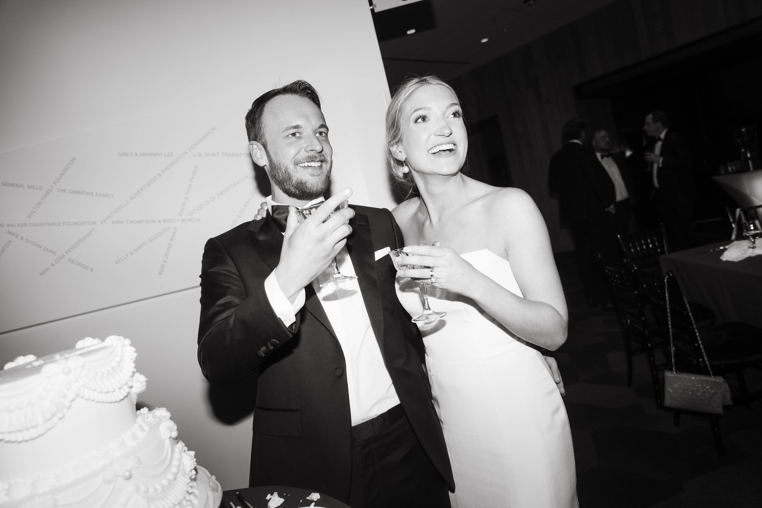 A couple in formal attire holding drinks and smiling at a wedding reception, with a wedding cake in the foreground and guests in the background. The setting appears to be indoors with elegant décor.